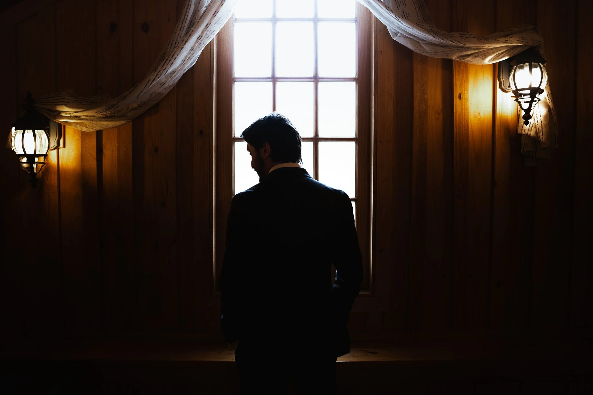 Man in a suit standing with hands in pockets, facing a window with light streaming in, in a wood-paneled room with two wall-mounted lamps.