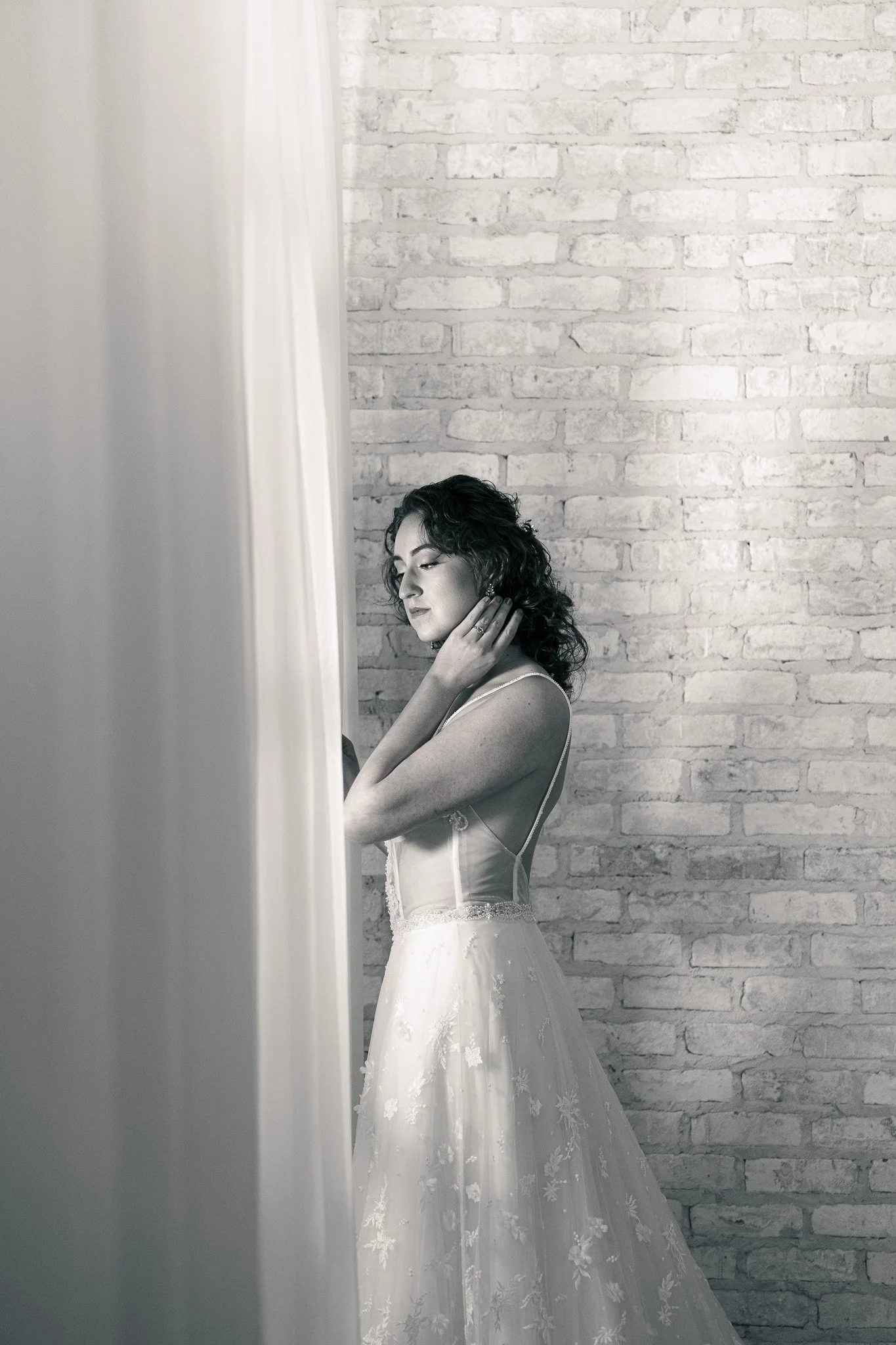 A woman in a wedding dress standing in front of a brick wall, partially covered by a curtain, touching her neck with her eyes closed.
