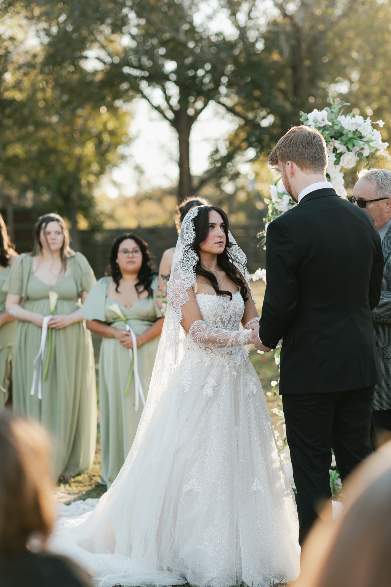 A bride and groom exchanging vows during an outdoor wedding ceremony with bridesmaids and guests in the background, surrounded by trees and warm sunlight.