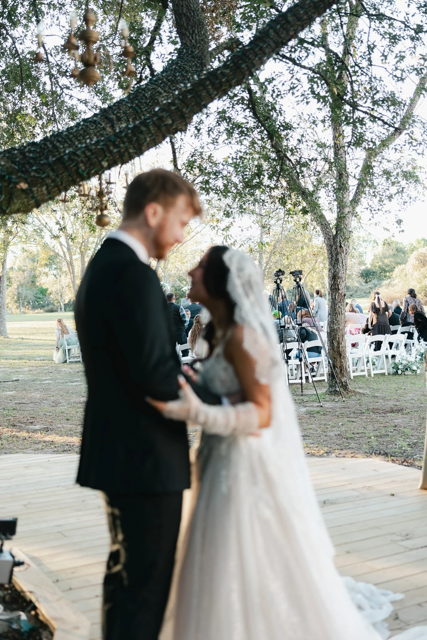 A newlywed couple sharing a kiss under a large tree during an outdoor wedding ceremony, with chairs and guests in the background.