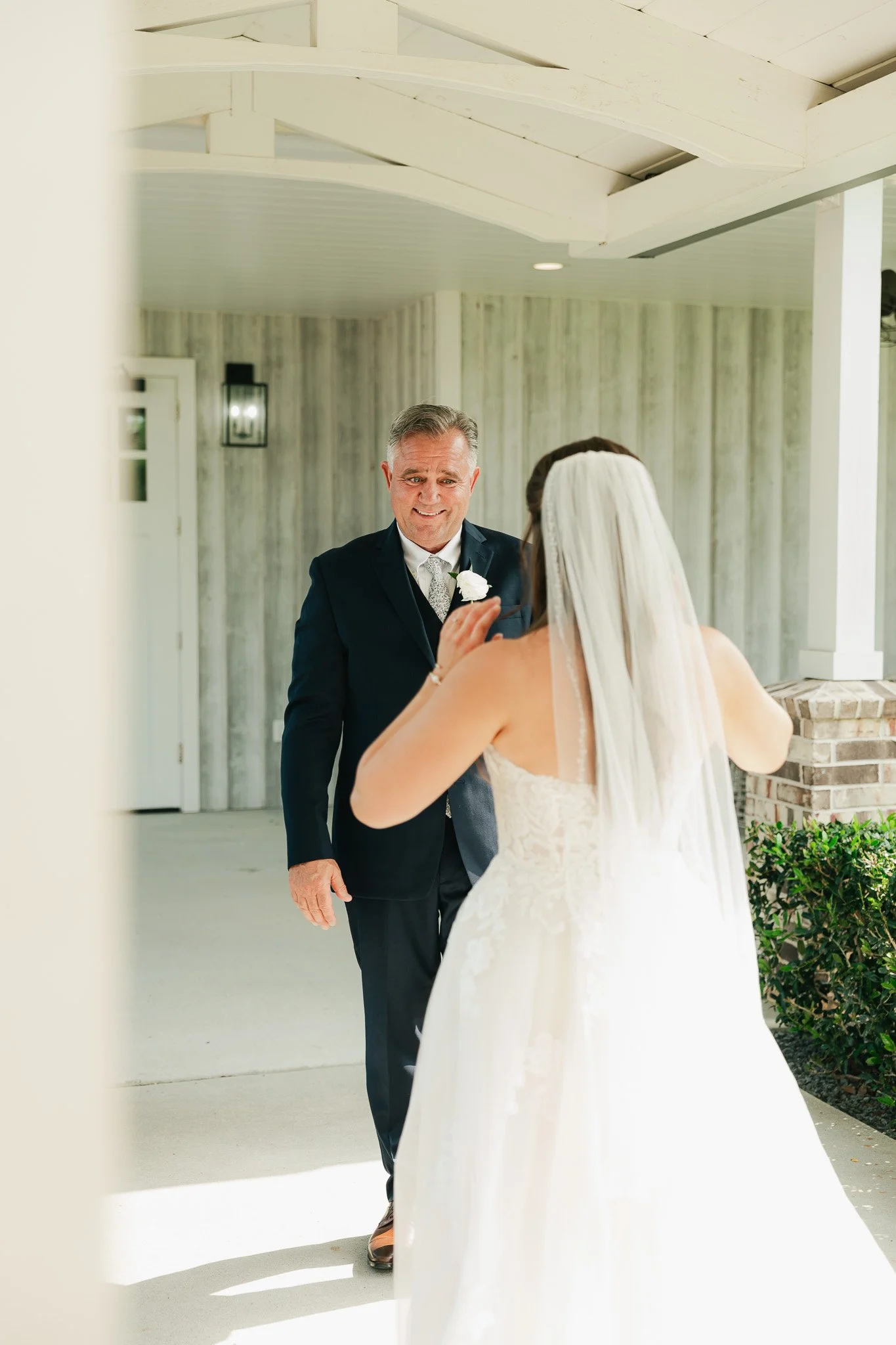 An elderly man in a black suit with a white boutonniere is smiling at a woman in a wedding dress and veil during a wedding moment outside a light-colored building.