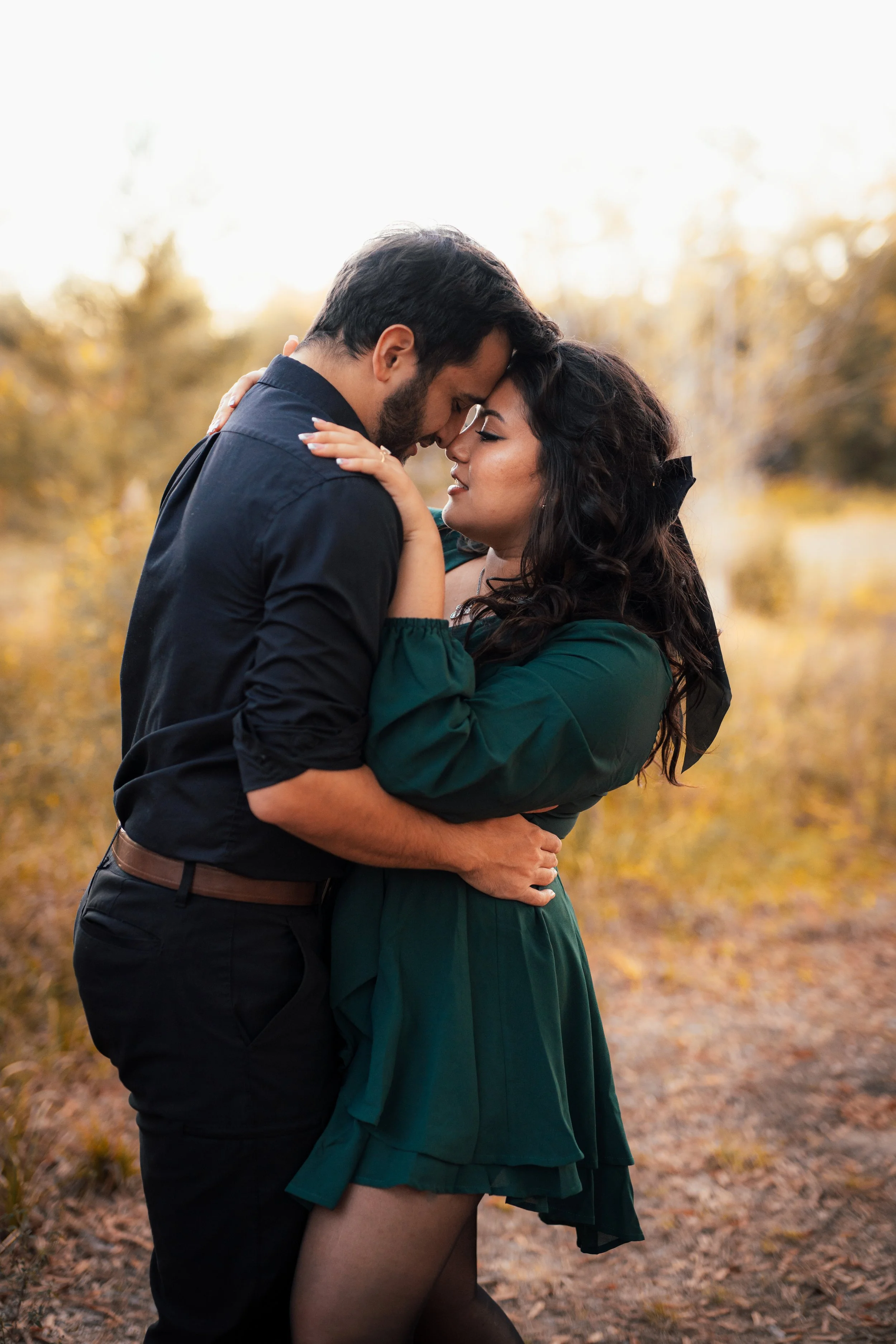 A couple embraces and touches foreheads in an outdoor setting during golden hour, surrounded by autumn trees.