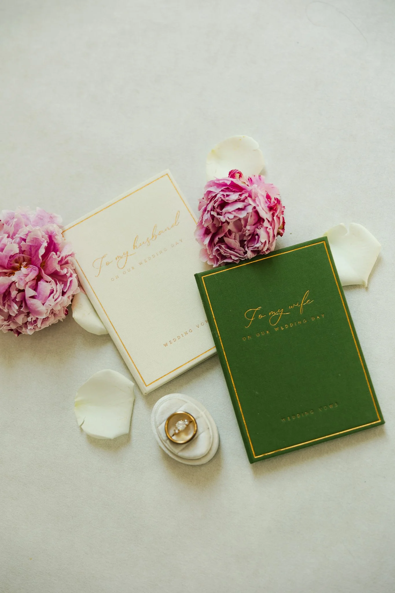 Wedding invitation cards, pink carnations, white rose petals, and a ring box with a gold wedding band inside, arranged on a white surface.