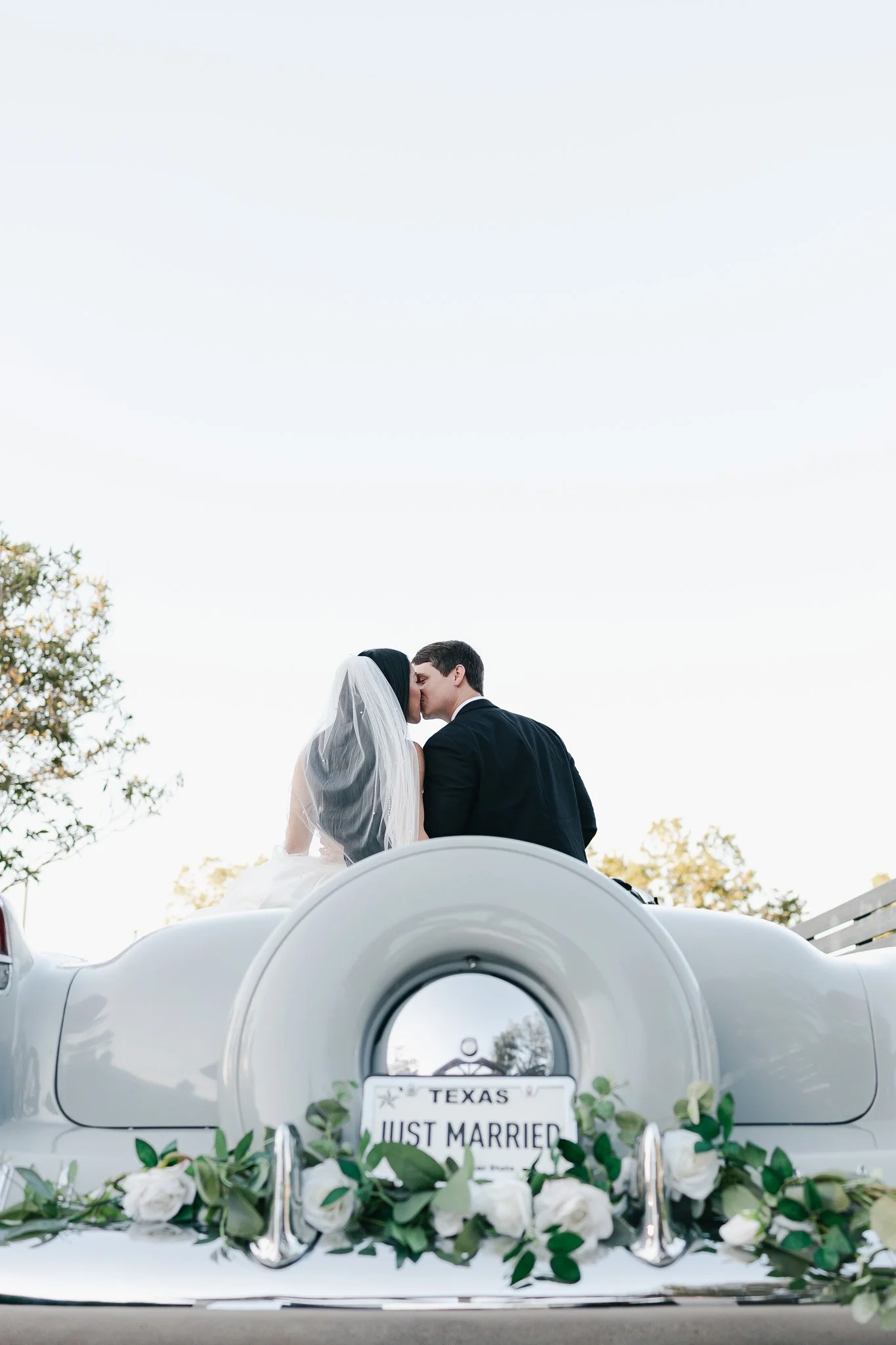 A newlywed couple kissing in a vintage car decorated with white roses and greenery, with a license plate that reads 'TEXAS JUST MARRIED,' and a clear sky and trees in the background.