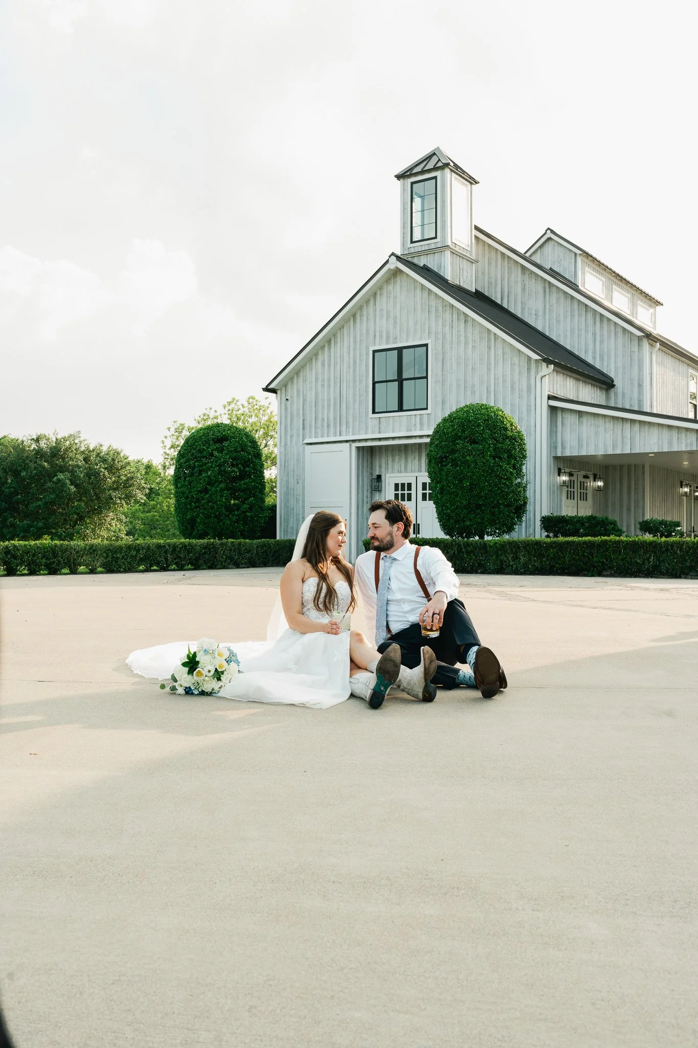 A couple dressed in wedding attire sitting on the ground in front of a large white barn-style house, holding drinks and looking at each other.