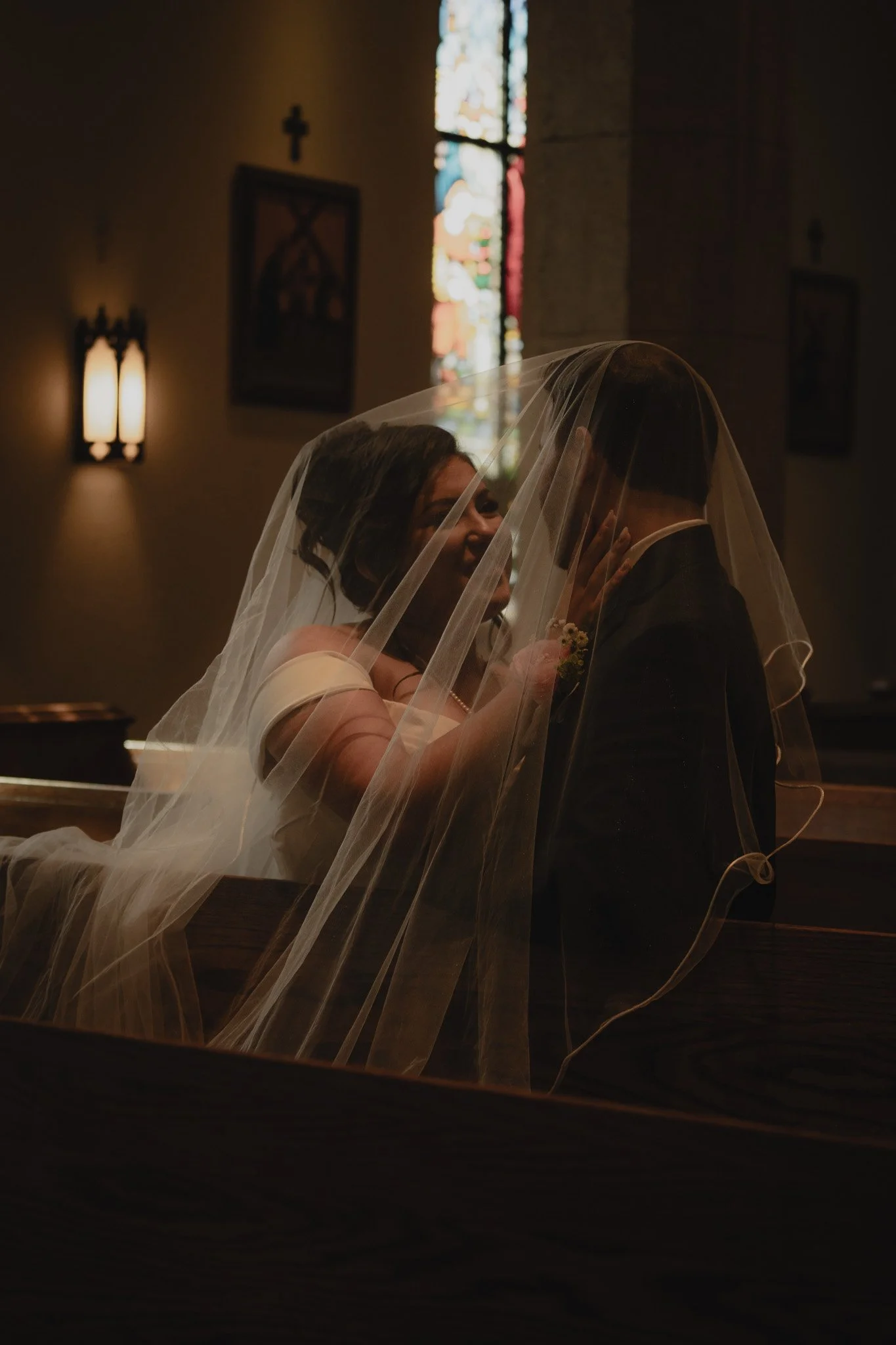 A bride and groom sharing a close, intimate moment under a wedding veil inside a church with stained glass windows.
