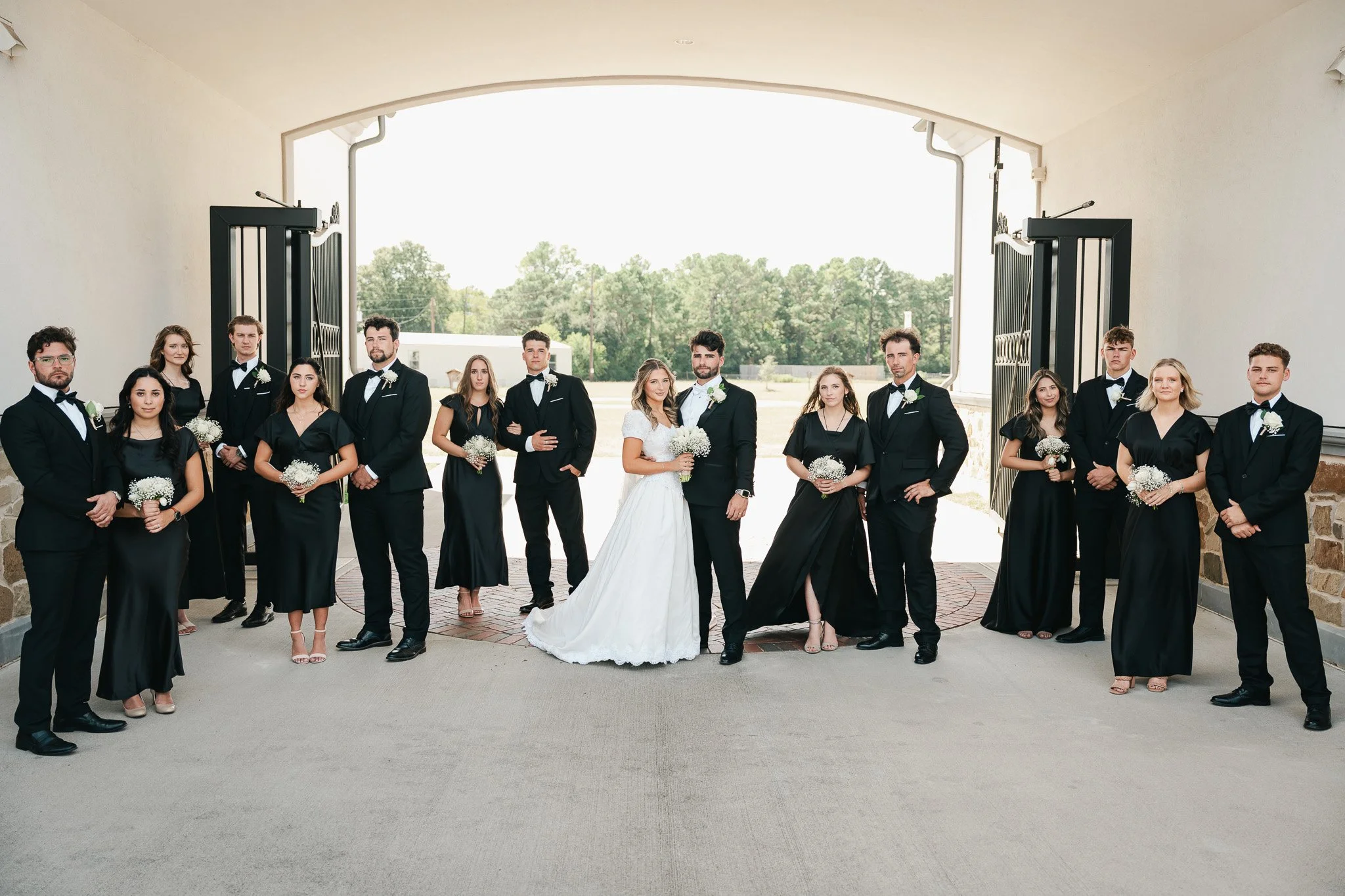 A wedding party with bridesmaids and groomsmen standing together outdoors under a large open gate. The bride and groom stand in the center, surrounded by their wedding party, all dressed in formal attire. The backdrop includes trees and a clear sky.