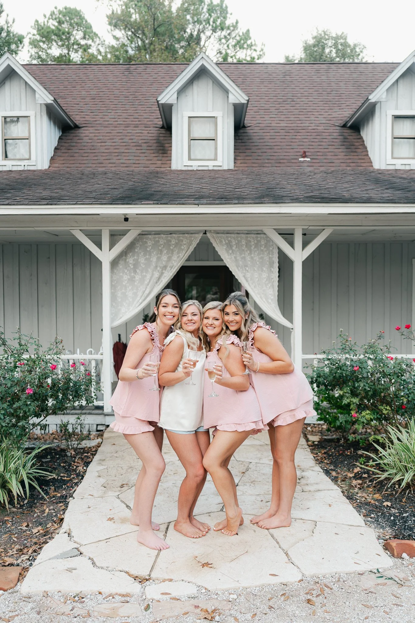 A group of five women standing together outdoors on a stone pathway, smiling and holding glasses, with a white house and garden in the background.