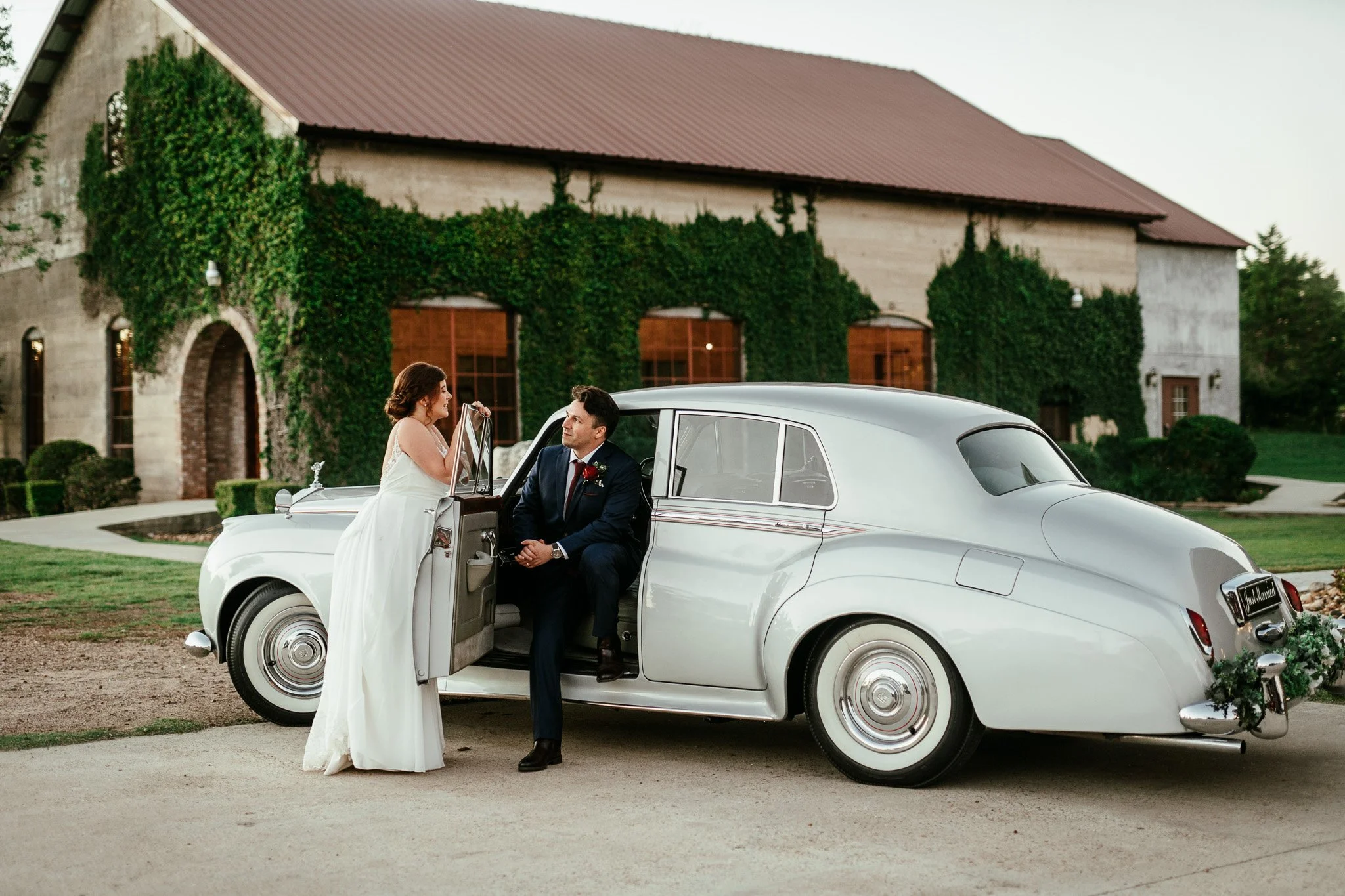 Bride in white dress and groom in dark suit sitting beside a vintage white car, outside a large building covered in green ivy.