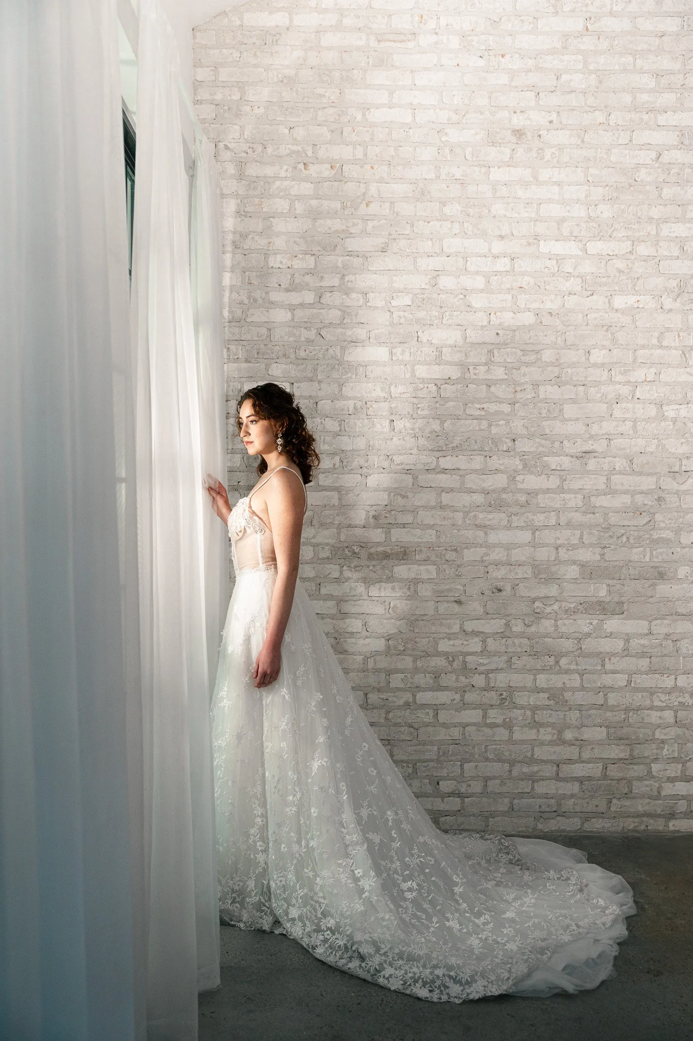 A woman in a wedding dress standing next to sheer white curtains, facing the window in a room with a white brick wall.