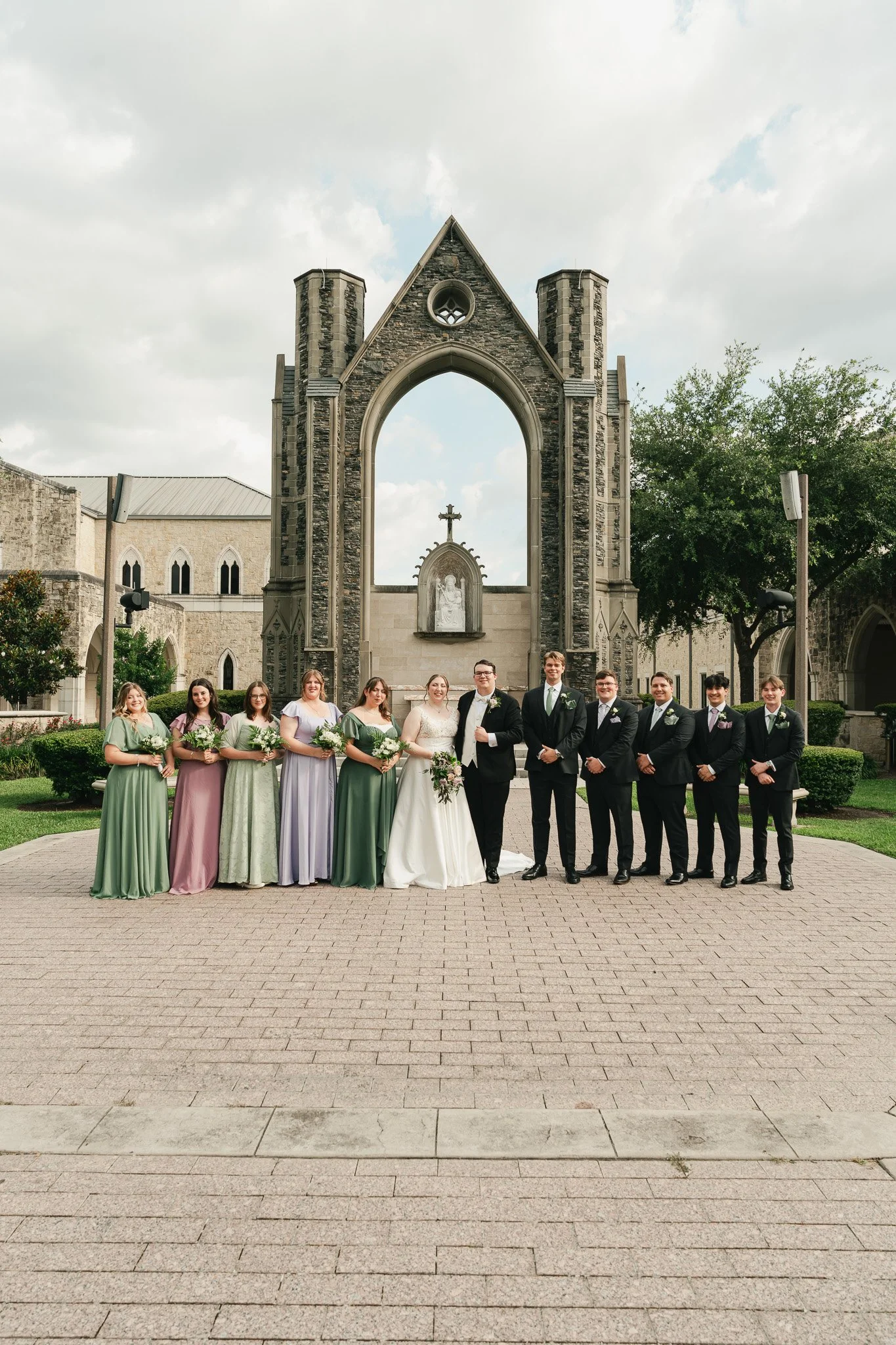 A wedding party of fourteen people, including a bride and groom, standing in front of a stone church during the daytime. The bride wears a white dress with a train, and the groom is in a black suit. Bridesmaids wear pastel-colored dresses, and grooms