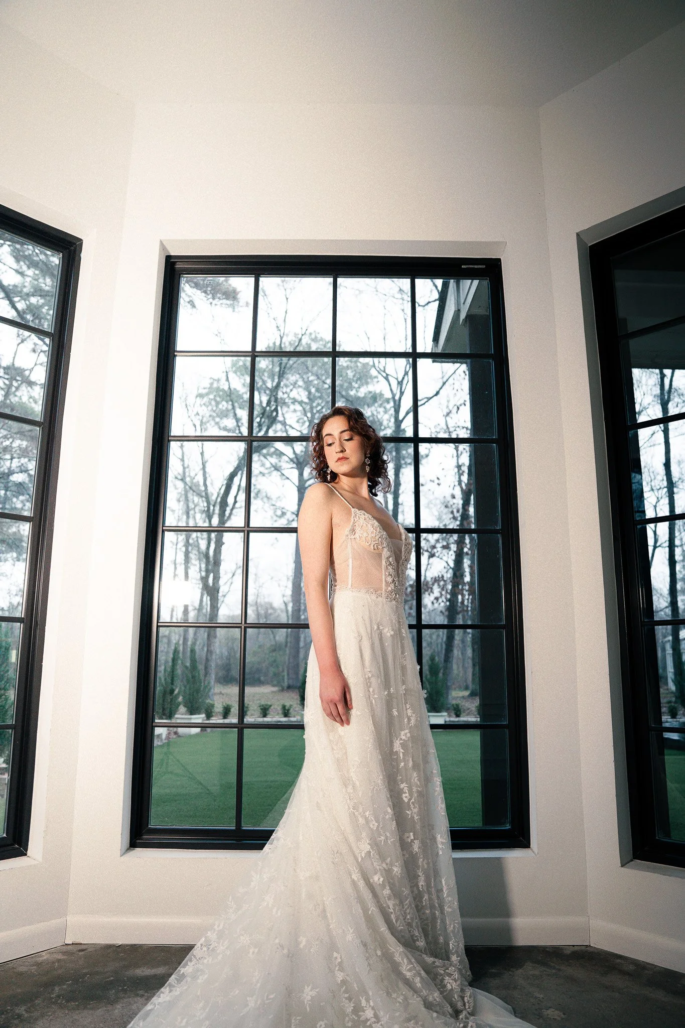 Bride in a white wedding gown standing inside a room with large windows and a view of trees outside.