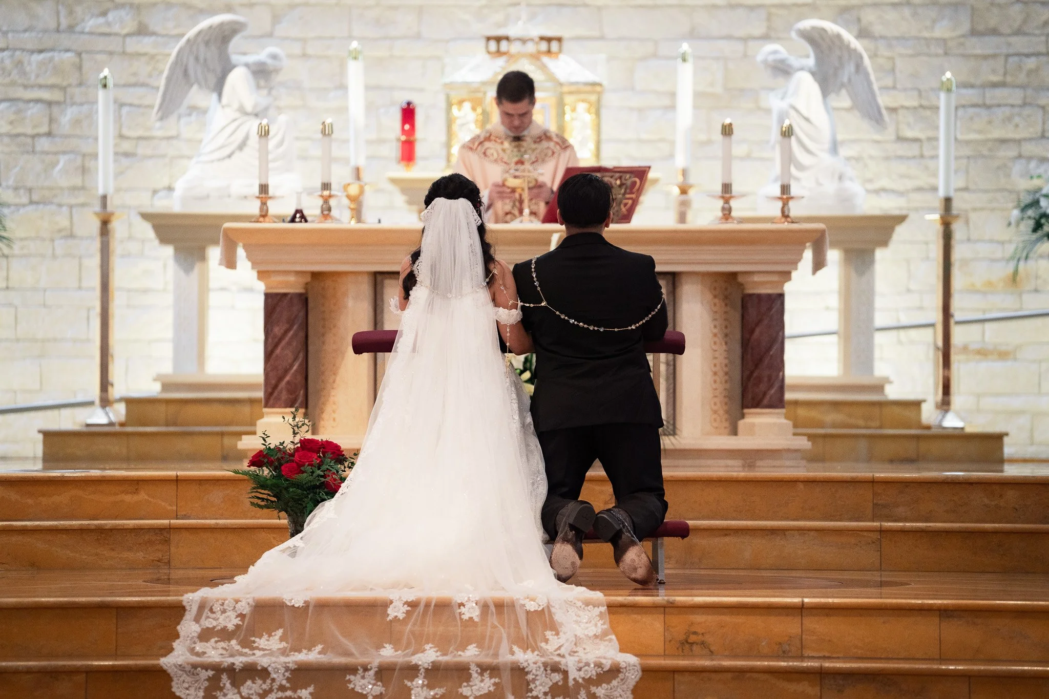 A bride and groom kneeling at the altar during a wedding ceremony inside a church, with a priest officiating. The bride is in a white wedding gown with a veil, and the groom is in a black suit decorated with a chain.