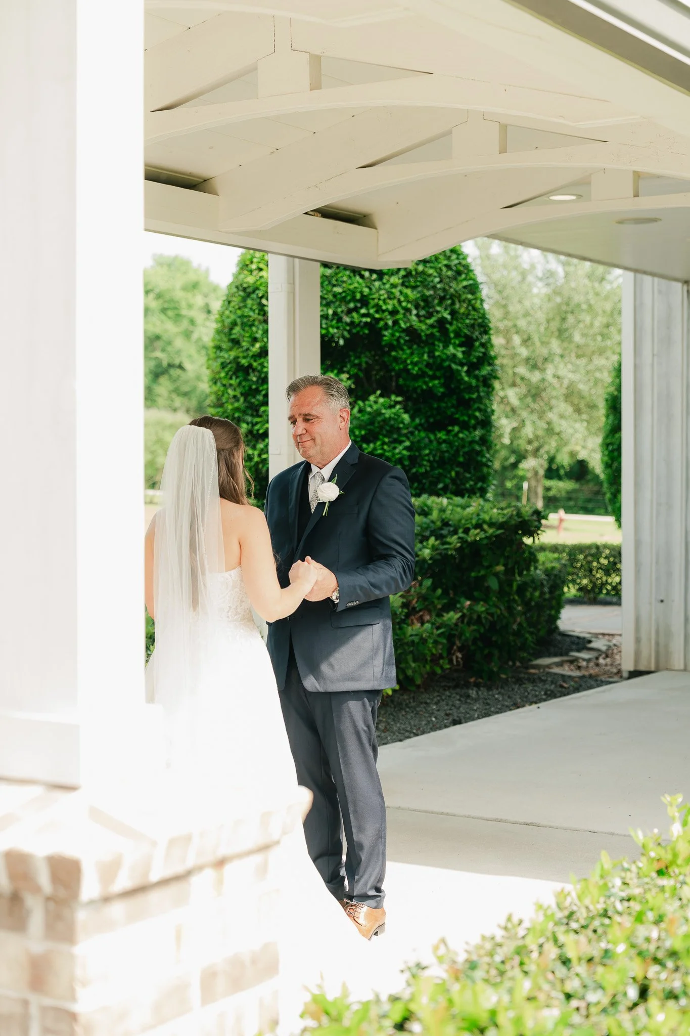A bride and groom holding hands during their wedding ceremony outdoors under a white pavilion with green trees in the background.