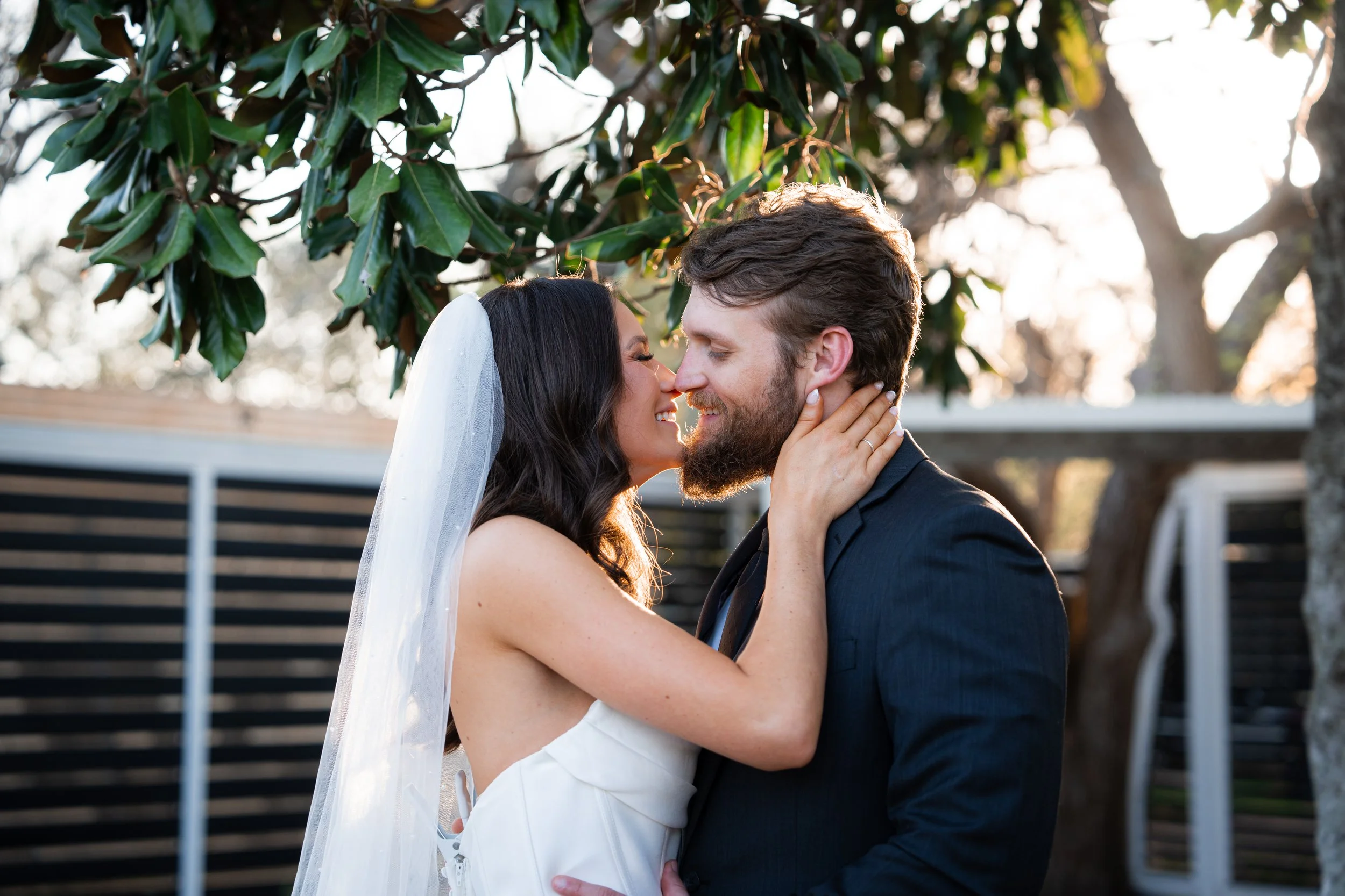 A bride and groom embrace outdoors during sunset, smiling and touching noses, with trees and a black fence in the background.