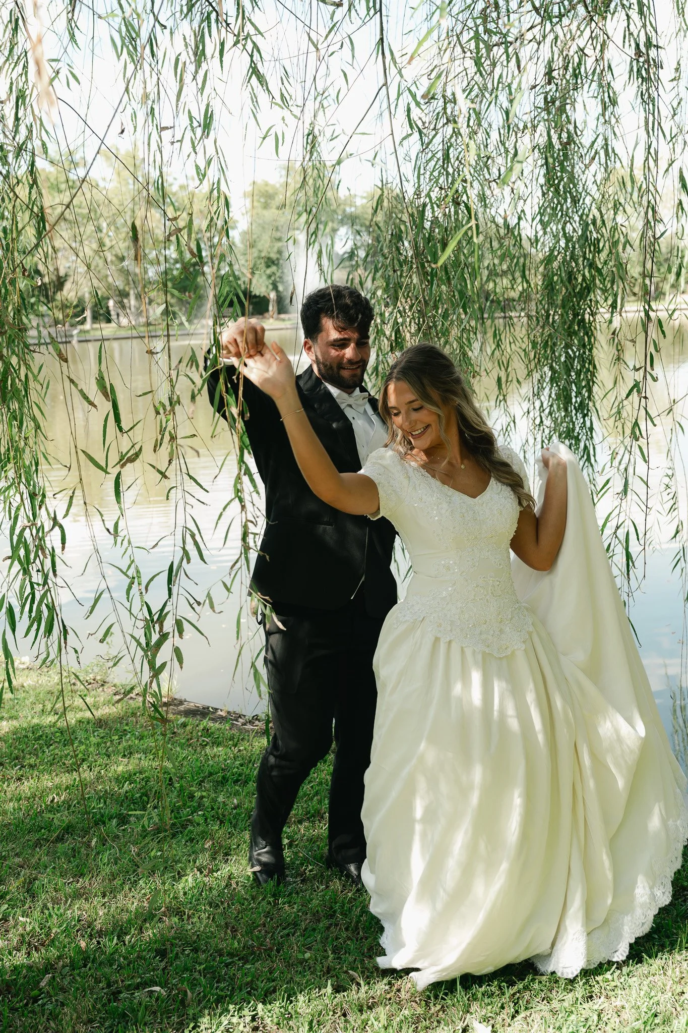 A newlywed couple dancing outdoors near a lake, surrounded by hanging willow branches and green grass.