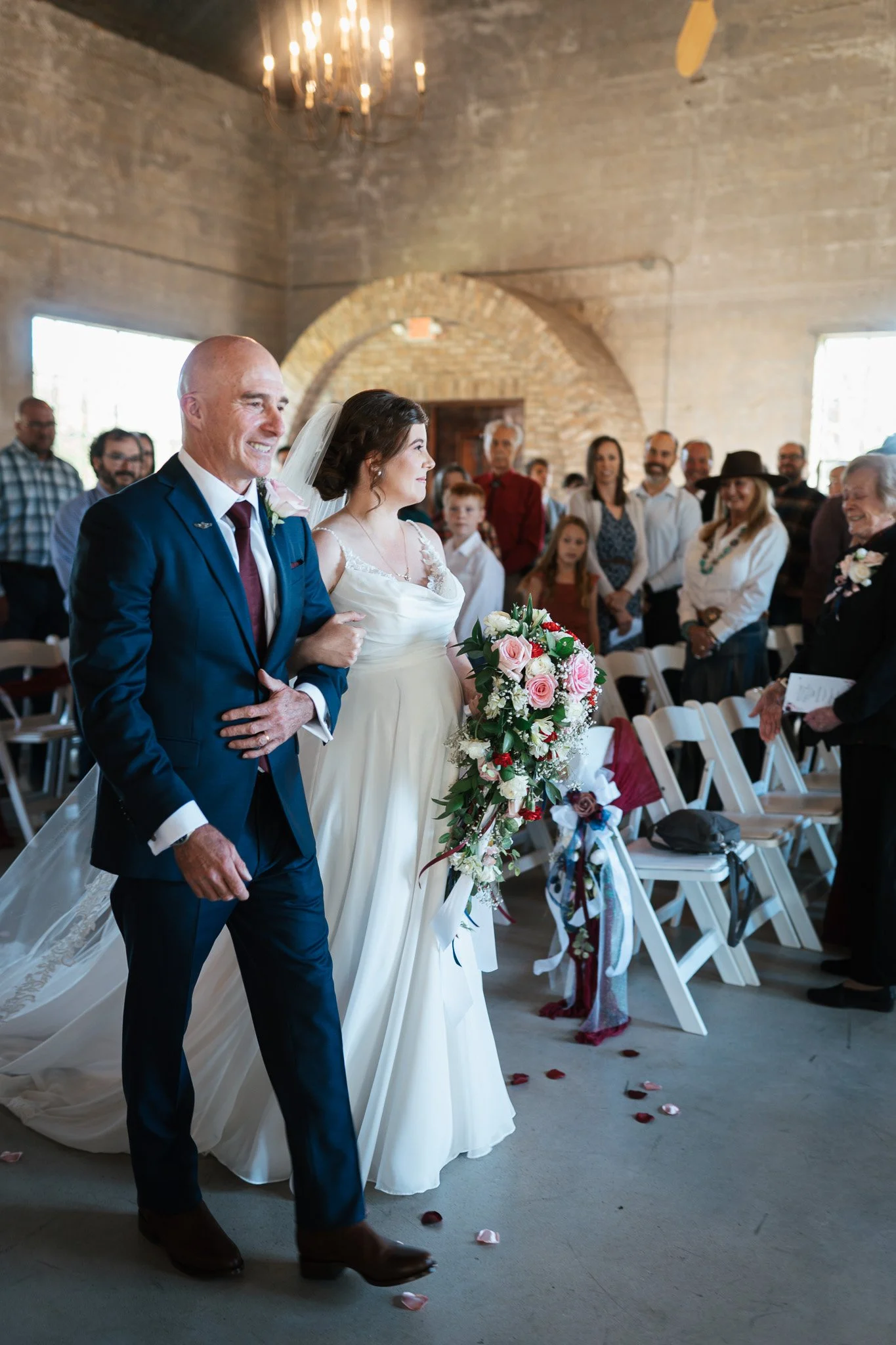 A bride walking down the aisle with her father at a wedding ceremony in a rustic indoor venue with stone walls, with guests standing and watching.