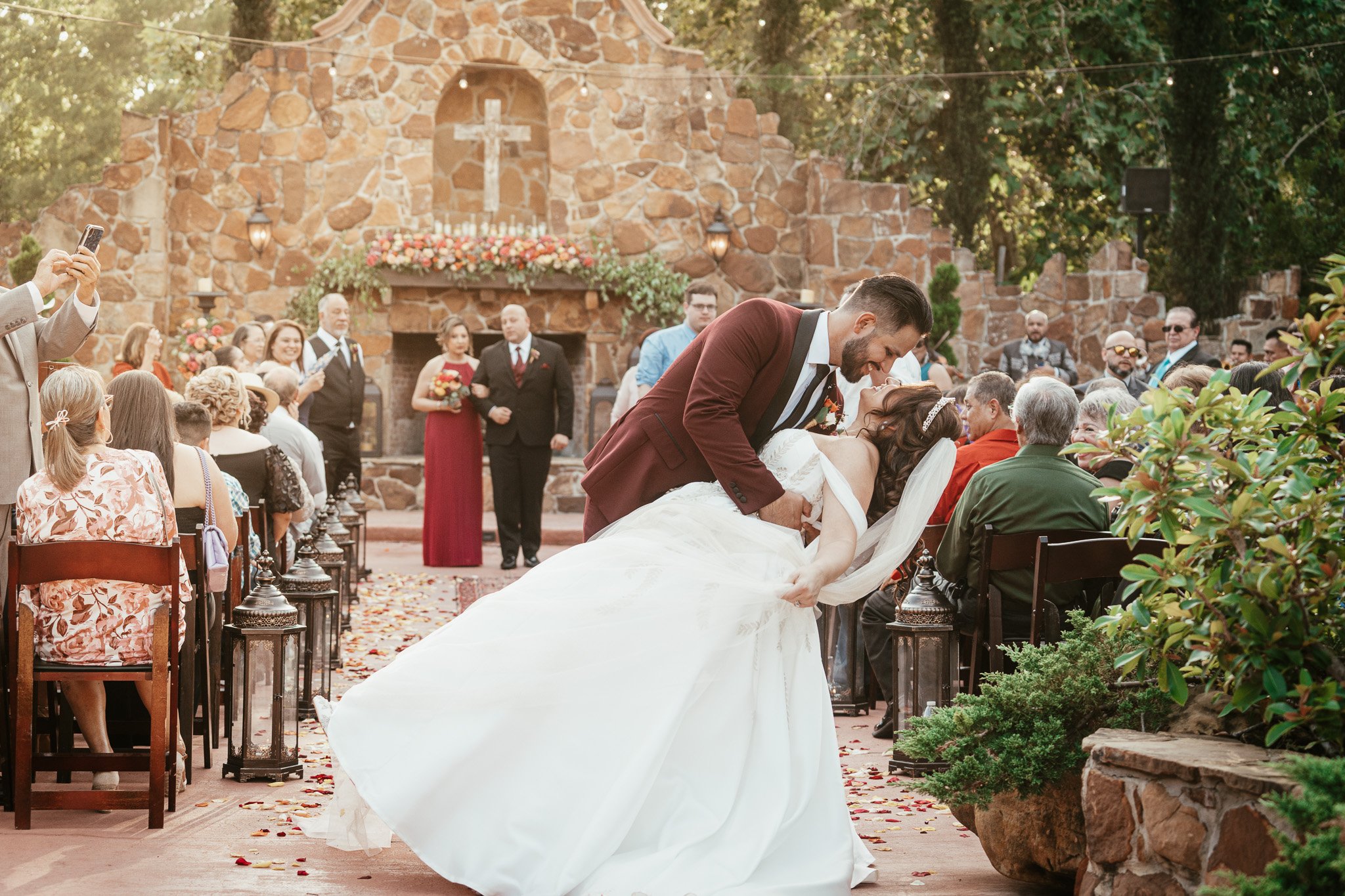 A newlywed couple sharing a kiss during their outdoor wedding ceremony in front of an altar with a cross, surrounded by guests seated on chairs, with some taking photos, and a stone backdrop decorated with flowers.