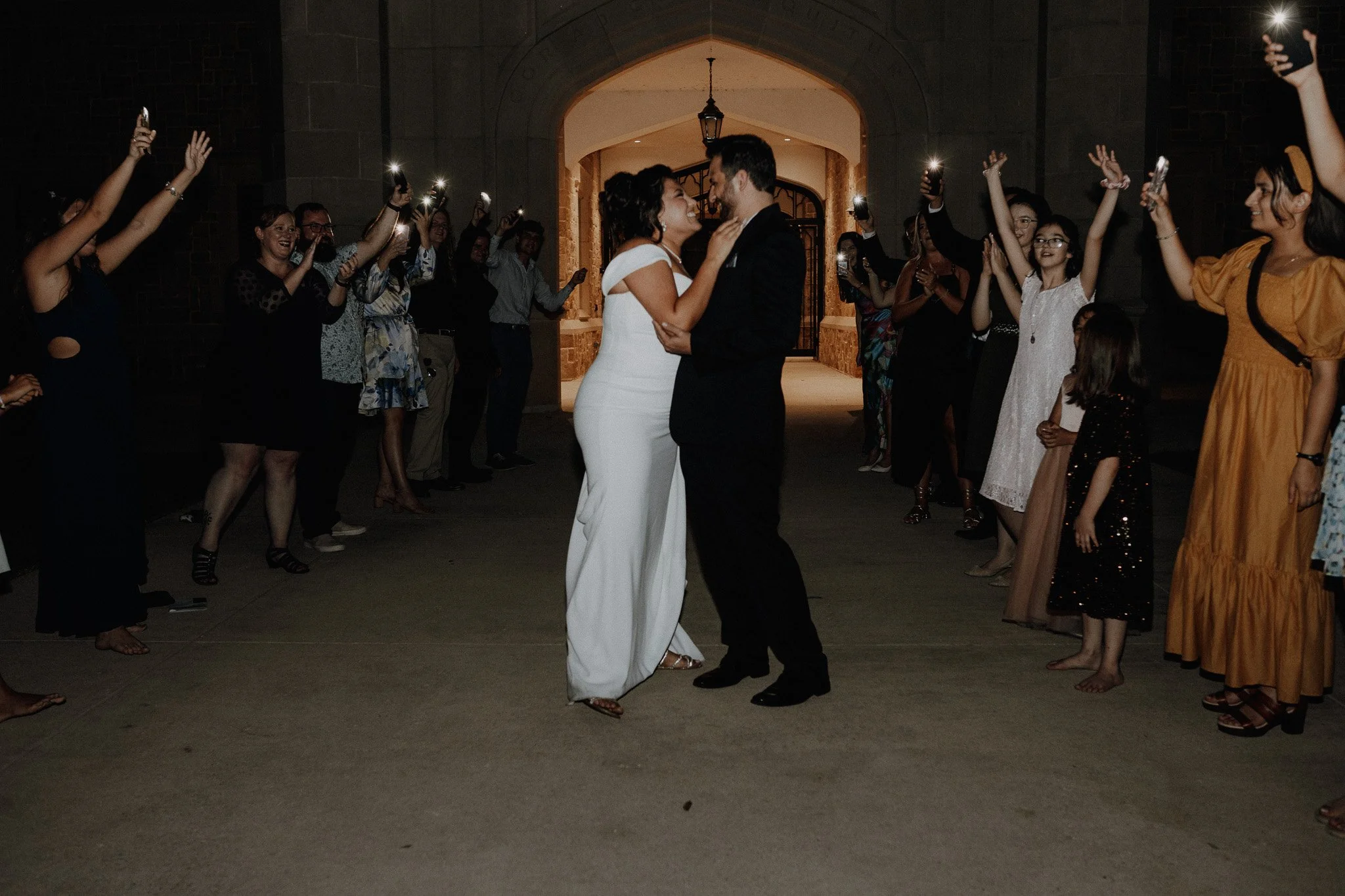 A bride and groom dancing in the center surrounded by guests at night, with many holding up phones capturing the moment, under an archway.