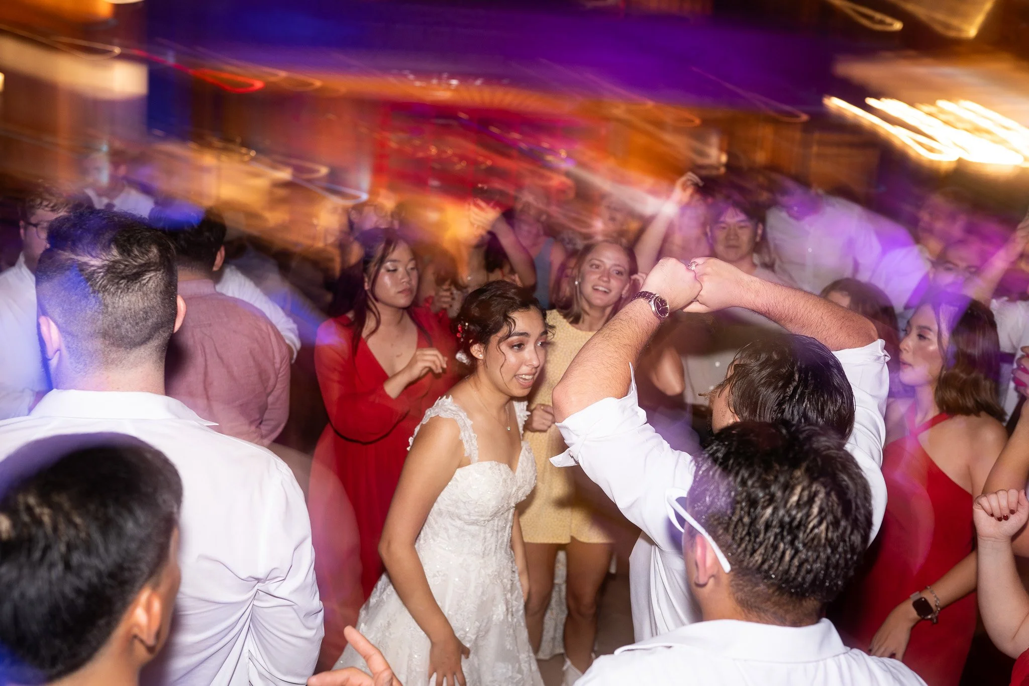 A bride in a white wedding dress dancing with a groom at a wedding reception, surrounded by guests dancing and enjoying the celebration.