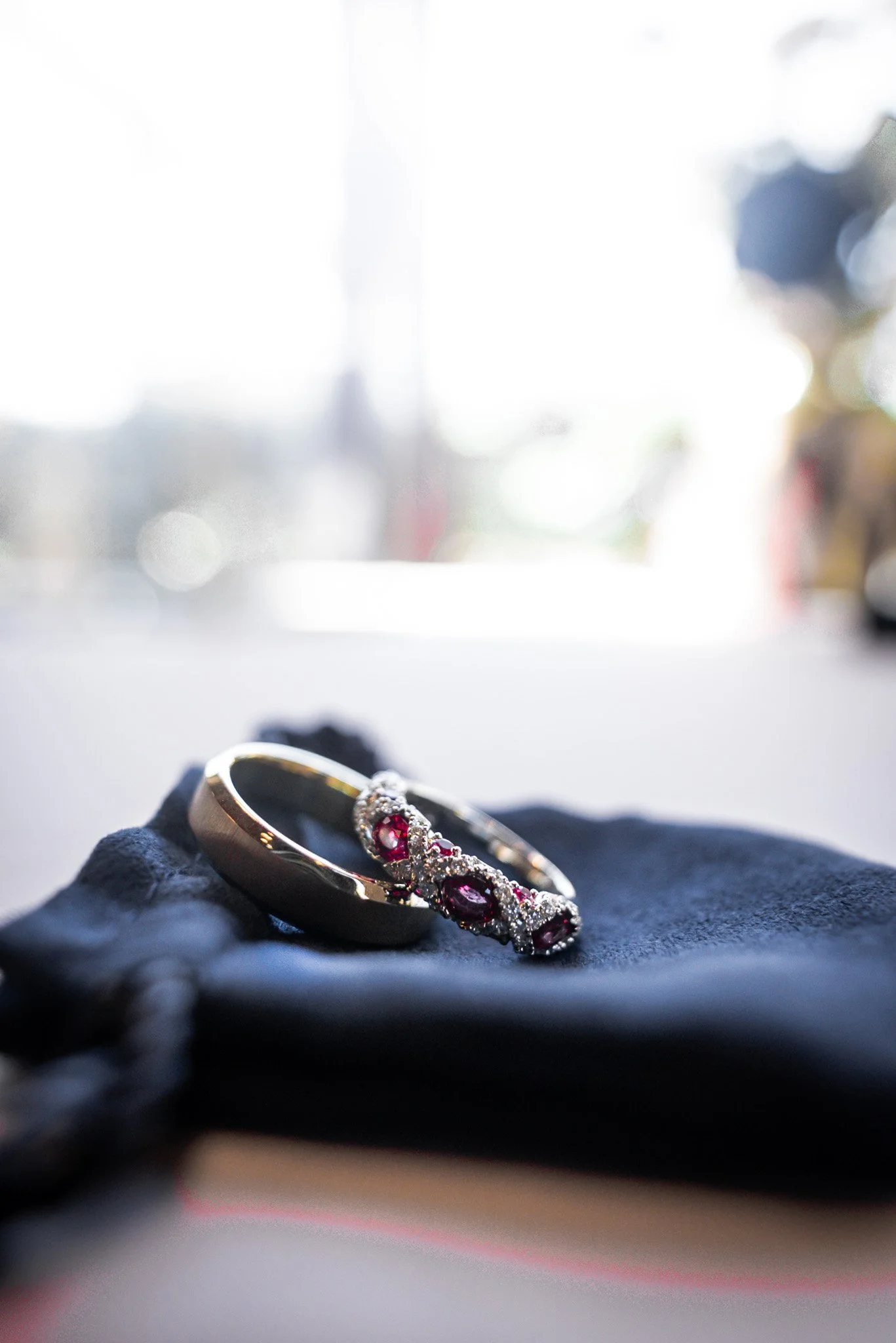 A gold ring and a gold bracelet with red gemstones and diamonds resting on a dark cloth with a bright, blurred window in the background.
