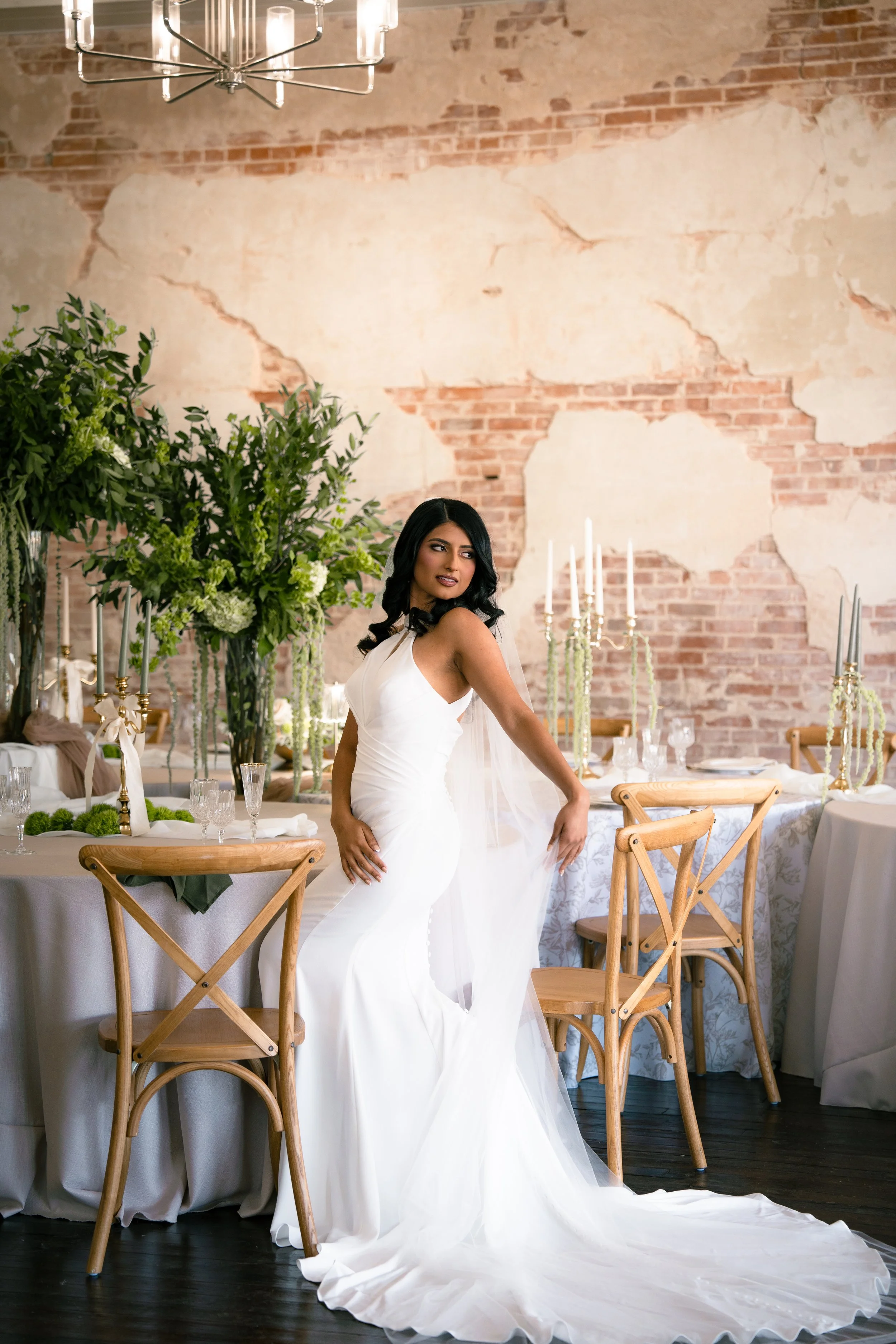 Woman in a white wedding dress posing in a decorated event space with a brick wall background and floral arrangements.