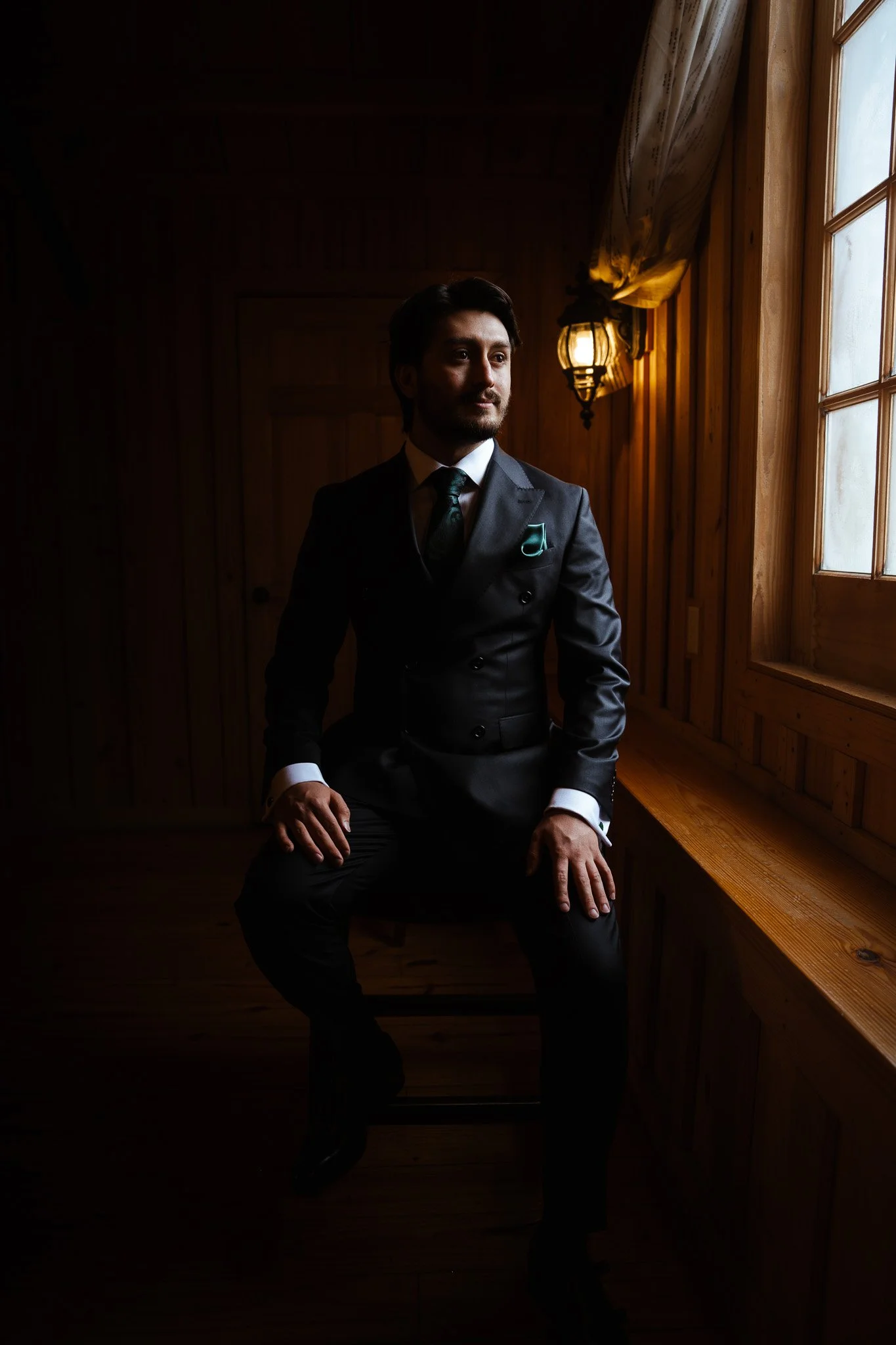 A man dressed in a dark formal suit sitting on a chair near a window in a wooden room, with sunlight coming through the window.