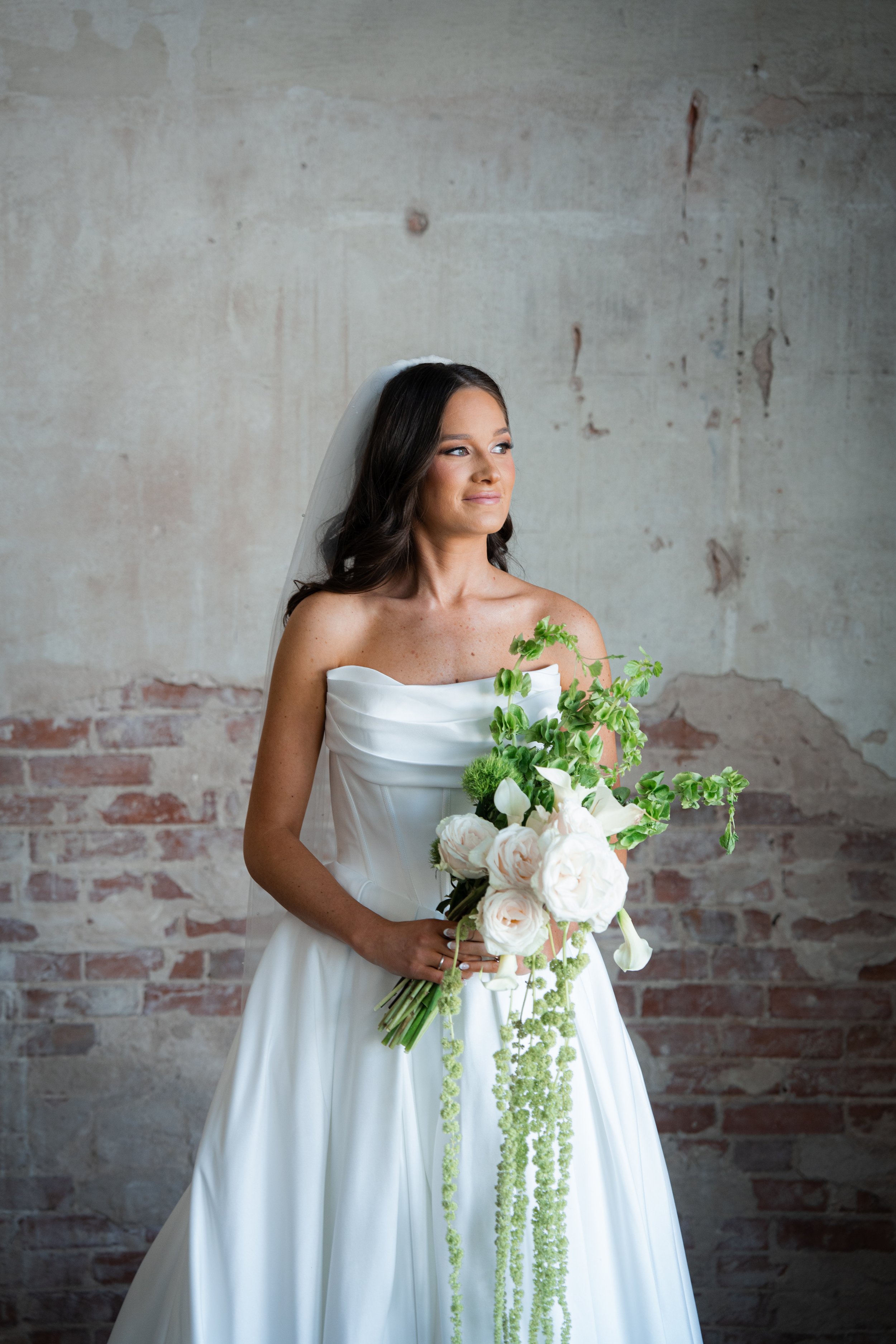Bride in a strapless white wedding dress holding a bouquet of white roses and greenery, standing against a textured brick wall.