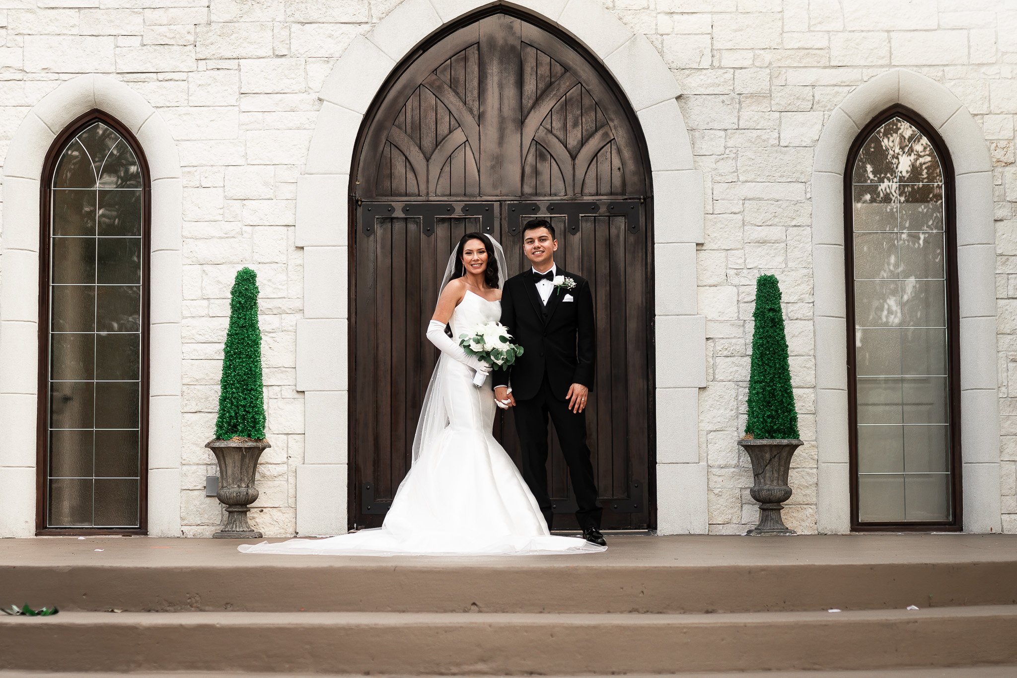 A bride and groom stand together outside a stone church, posing for a wedding photo. The bride is wearing a white wedding gown, gloves, and holding a bouquet, while the groom is dressed in a black tuxedo with a bow tie.