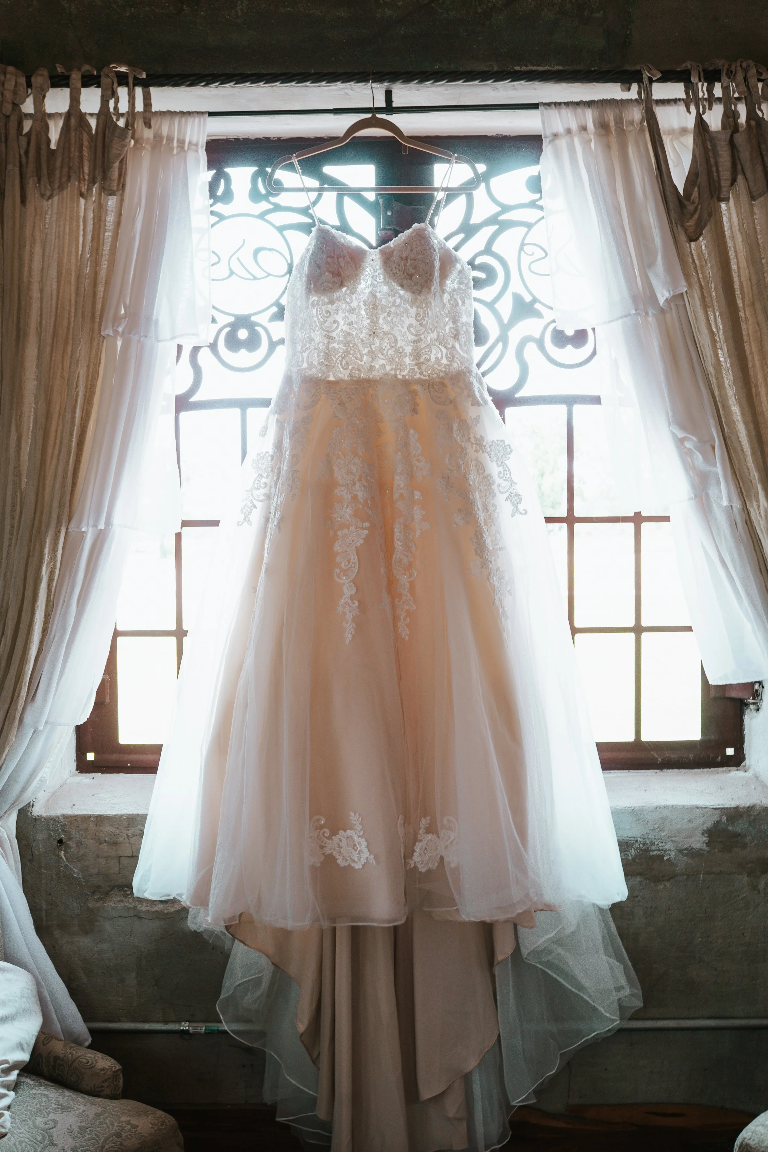 A wedding dress hanging on a hanger in front of a window with decorative ironwork, surrounded by curtains.
