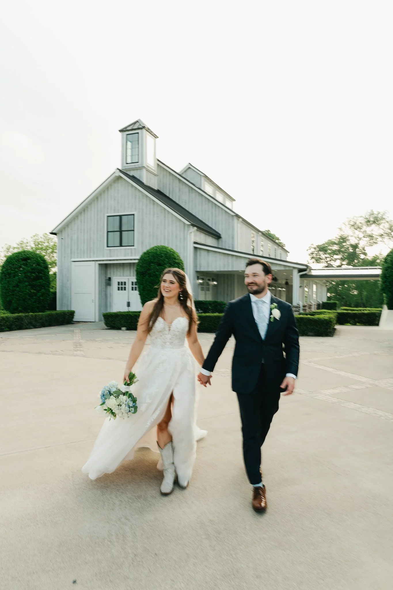 A bride and groom walking hand in hand outdoors in front of a white building with a small tower. The bride is in a white wedding dress holding a bouquet, and the groom is dressed in a navy suit with a boutonniere. They are smiling and appear happy.