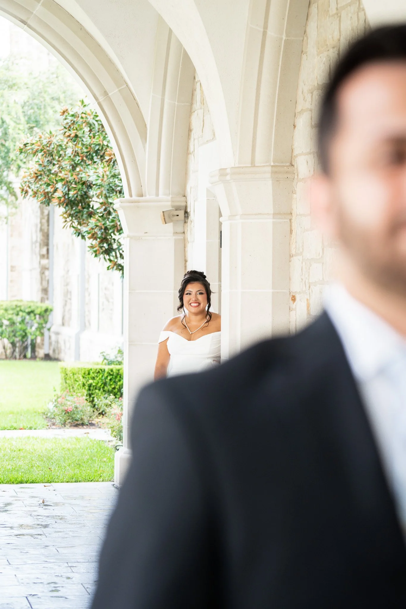 A bride in a white wedding dress smiling as she looks at a groom in a black tuxedo, who is blurred in the foreground, outside a stone archway with greenery and a tree in the background.