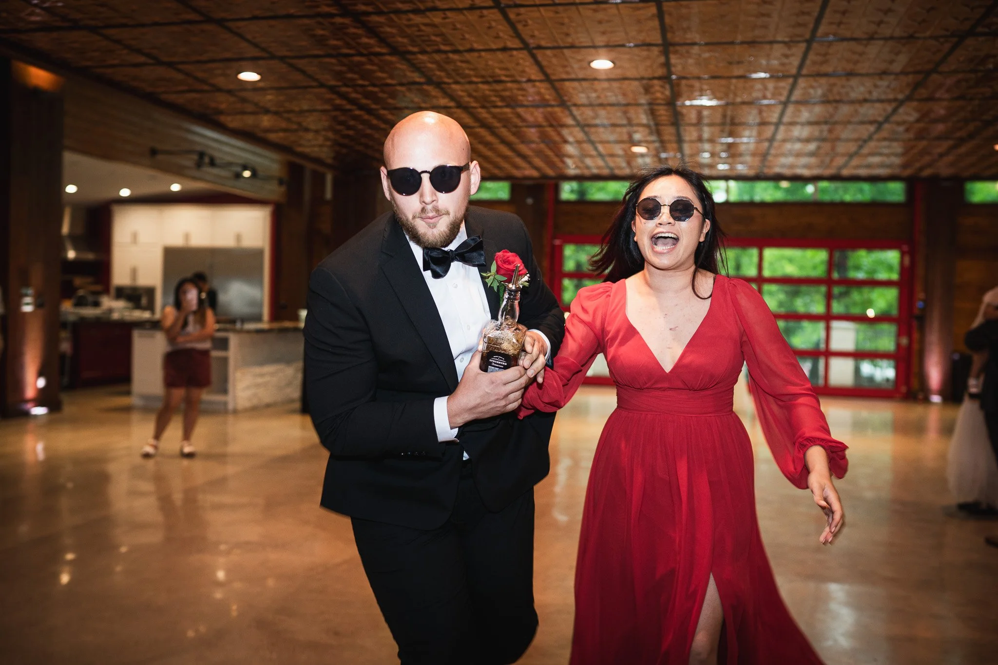 Man in a tuxedo and woman in a red dress wearing sunglasses, both smiling and holding a bottle with a red rose on top, in a spacious indoor setting with high ceiling and large windows.
