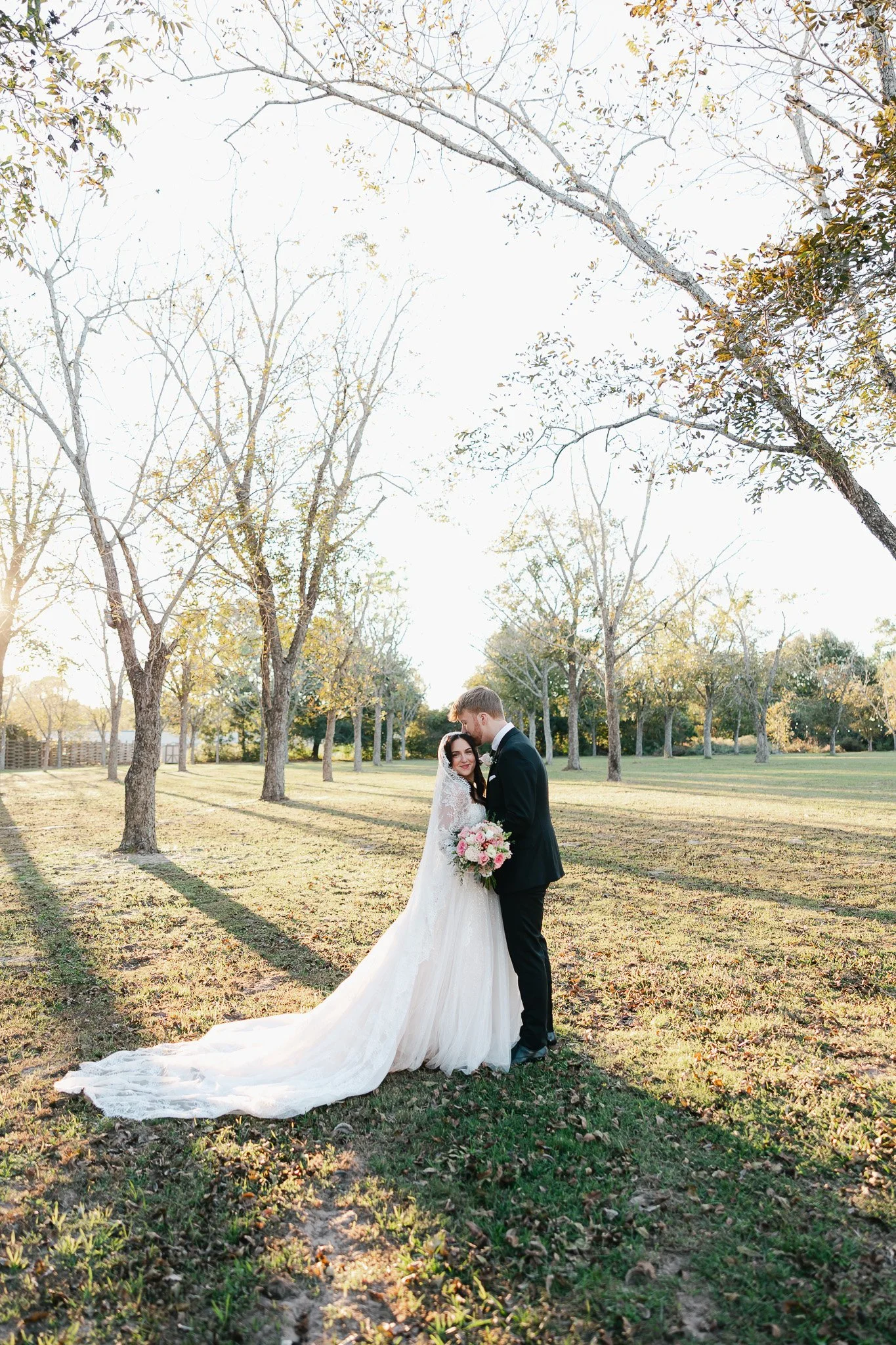 A bride and groom are standing close together in a park with trees and grass, holding each other and smiling. The bride is wearing a long white wedding gown and veil, and holding a bouquet of pink and white flowers. The groom is dressed in a black su