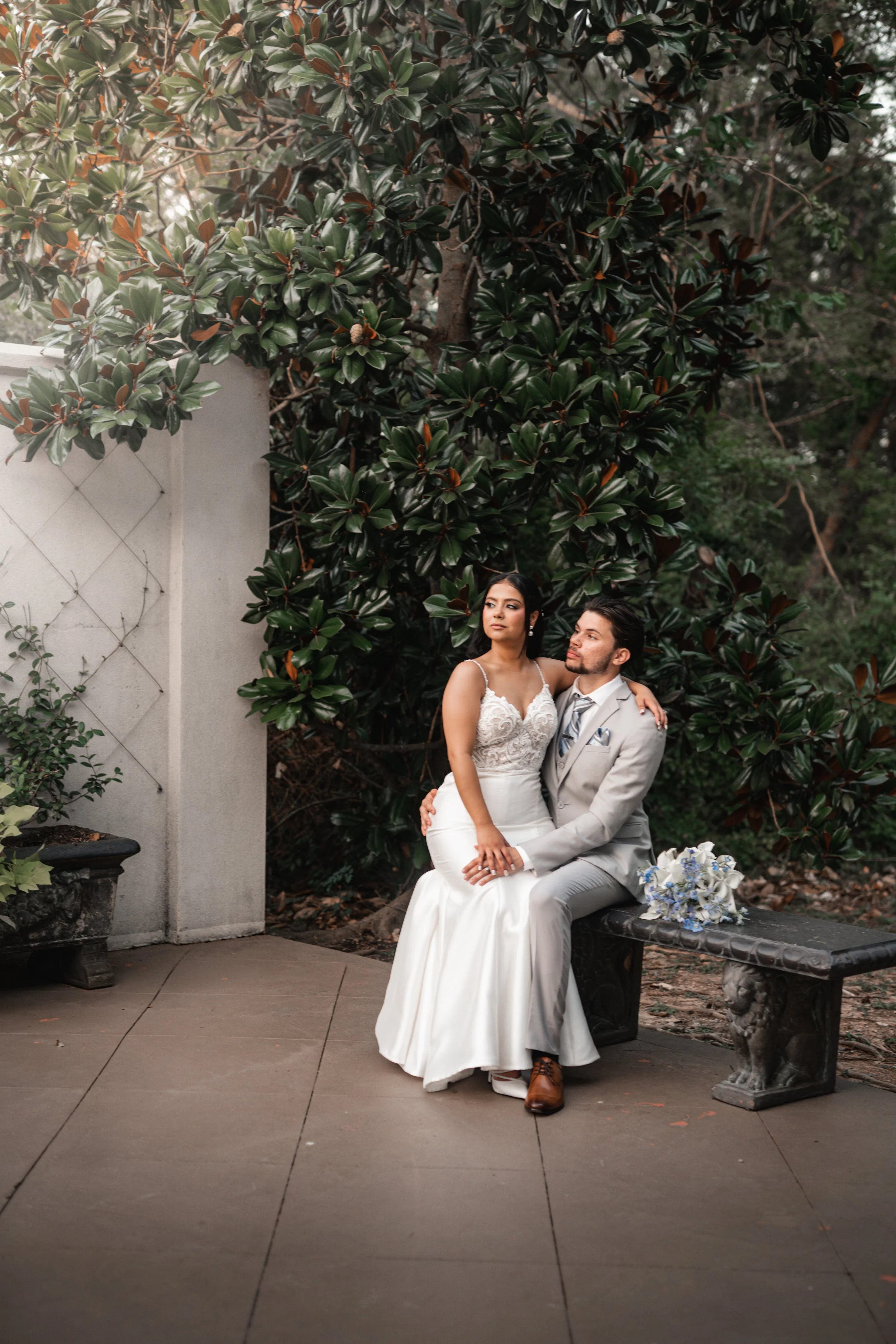 A bride seated on a bench with her groom standing next to her, outdoors with large green foliage in the background. The bride wears a white wedding dress and the groom wears a light gray suit. There is a bouquet of white and blue flowers on the bench