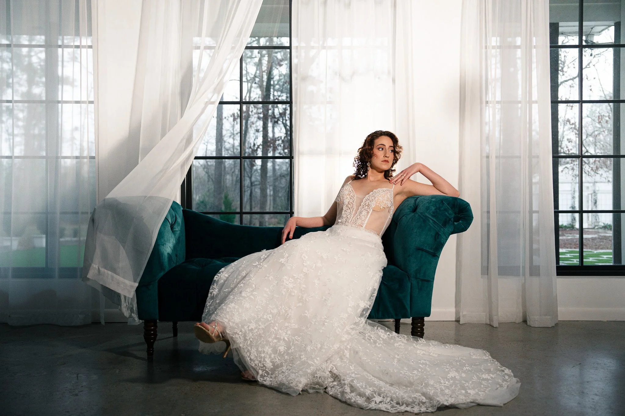 A woman in a white wedding dress sitting on a teal velvet sofa in front of large windows with sheer white curtains.