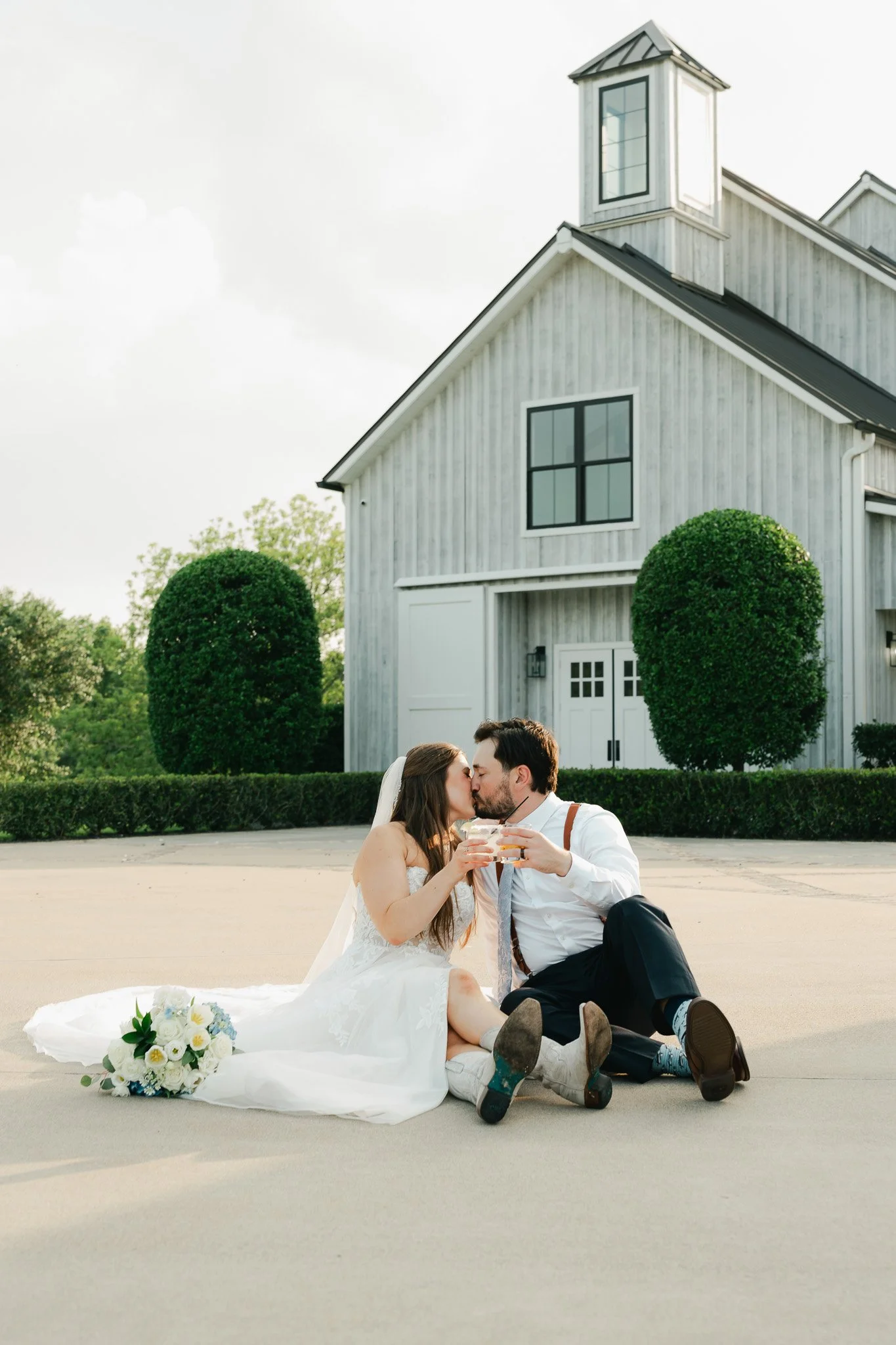 A bride and groom sitting on the ground in front of a white barn, kissing, holding glasses of champagne, with a bouquet resting on the bride's white wedding dress.