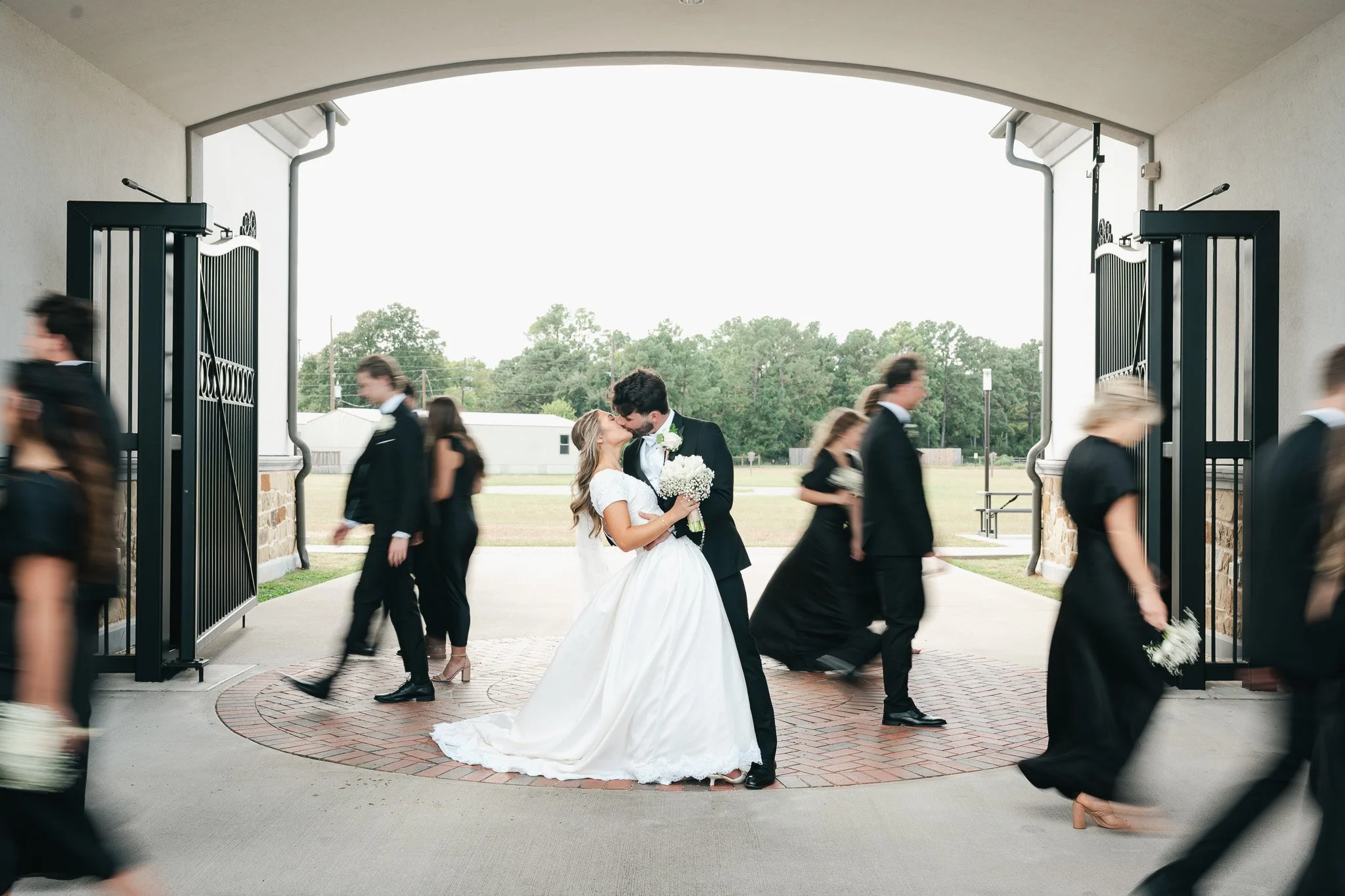 A bride and groom kiss under a large archway as wedding guests walk by, some blurry due to motion, during an outdoor wedding ceremony.