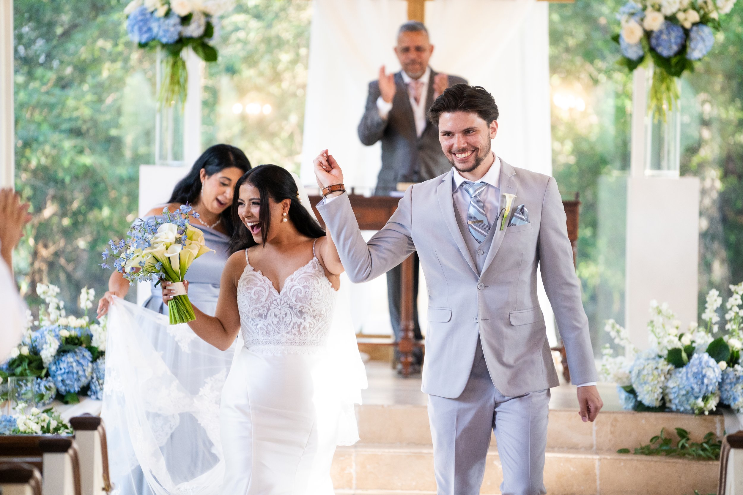 A joyful newlywed couple walking down the aisle in a wedding ceremony, surrounded by flowers, with the officiant clapping in the background.