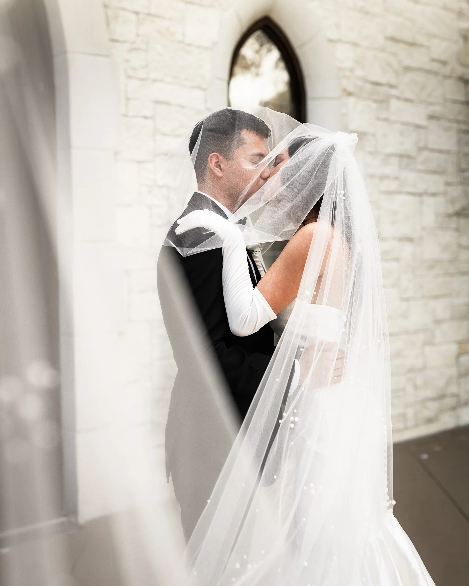 Bride and groom sharing a kiss behind a sheer wedding veil in a church with white brick walls.