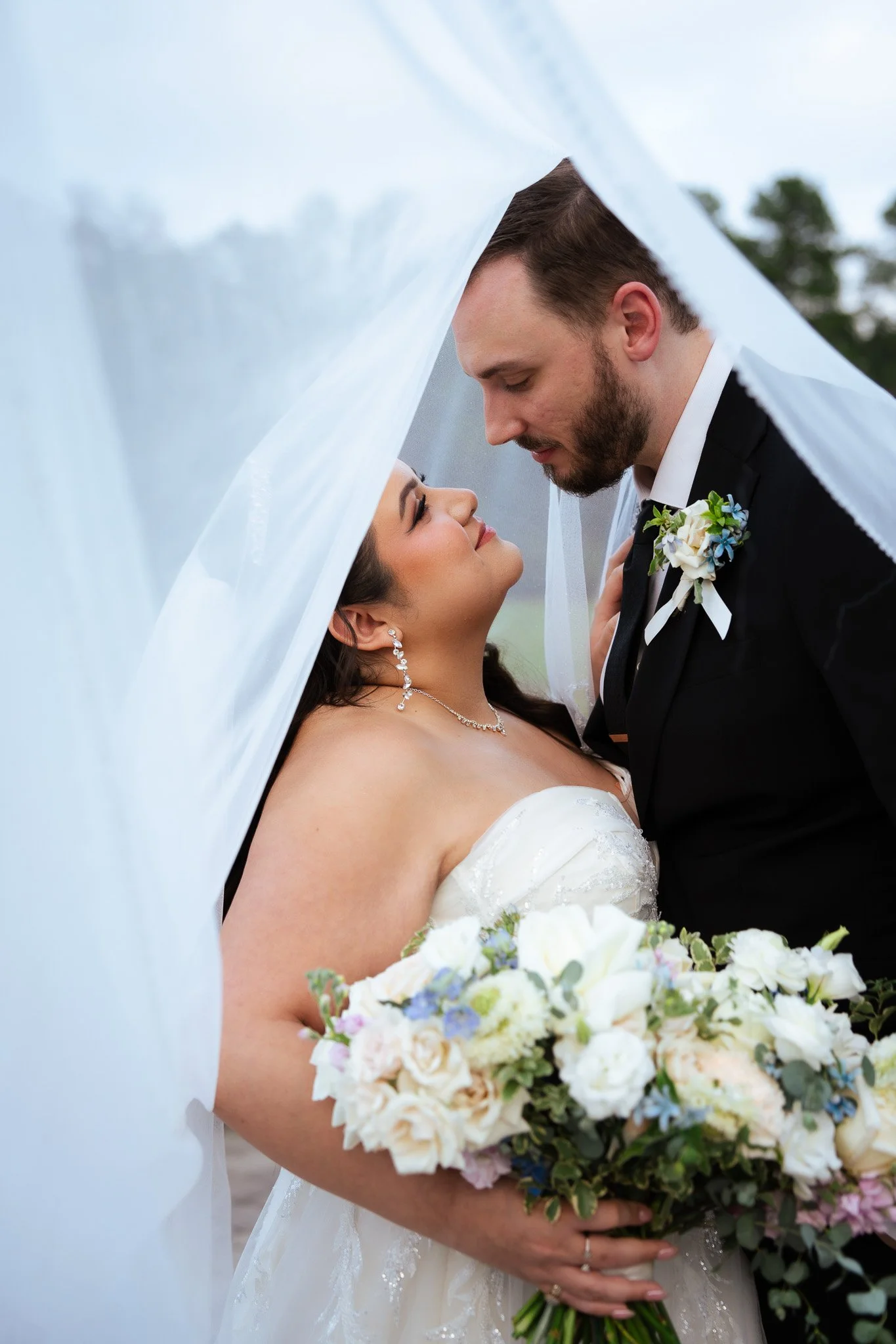 A bride and groom are close together under a white veil during their wedding, with the bride holding a bouquet of white and pastel flowers.