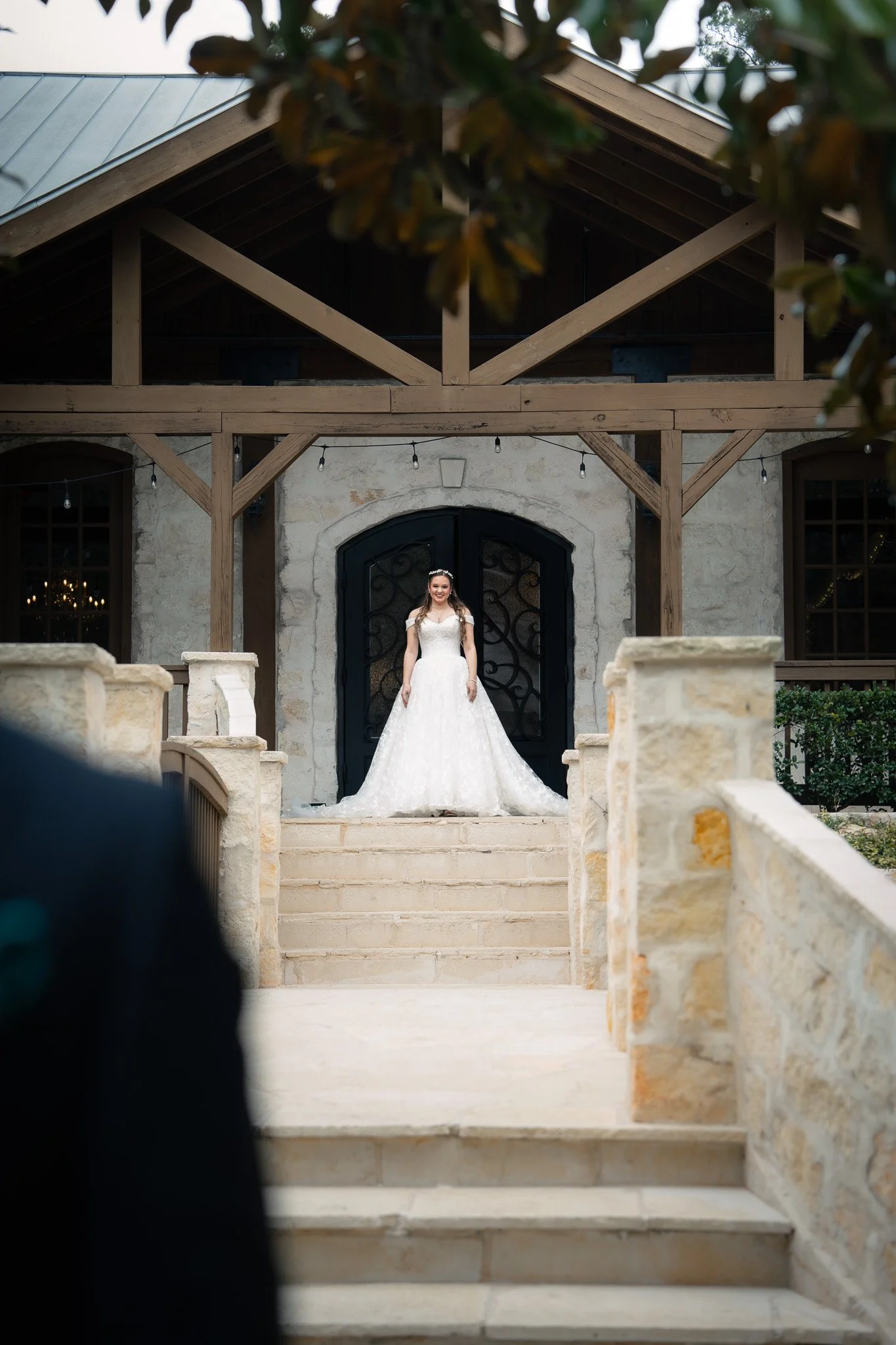 A bride in a white wedding dress stands on the steps in front of a solid black door with decorative ironwork at a wedding venue.
