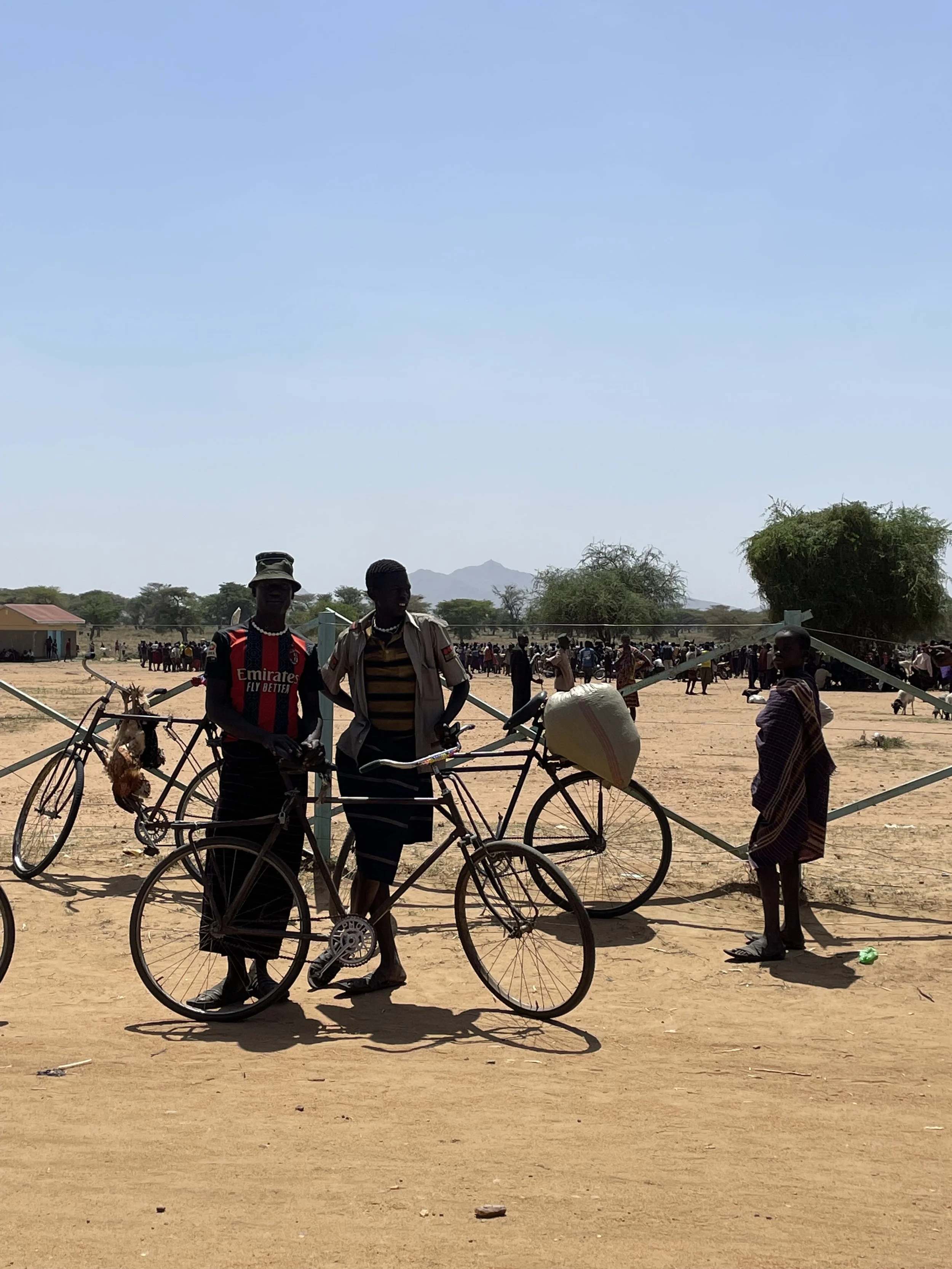 Two men with bicycles standing on a dirt ground in a rural area, with a woman standing nearby and a crowd in the background under a blue sky in Eastern Uganda.