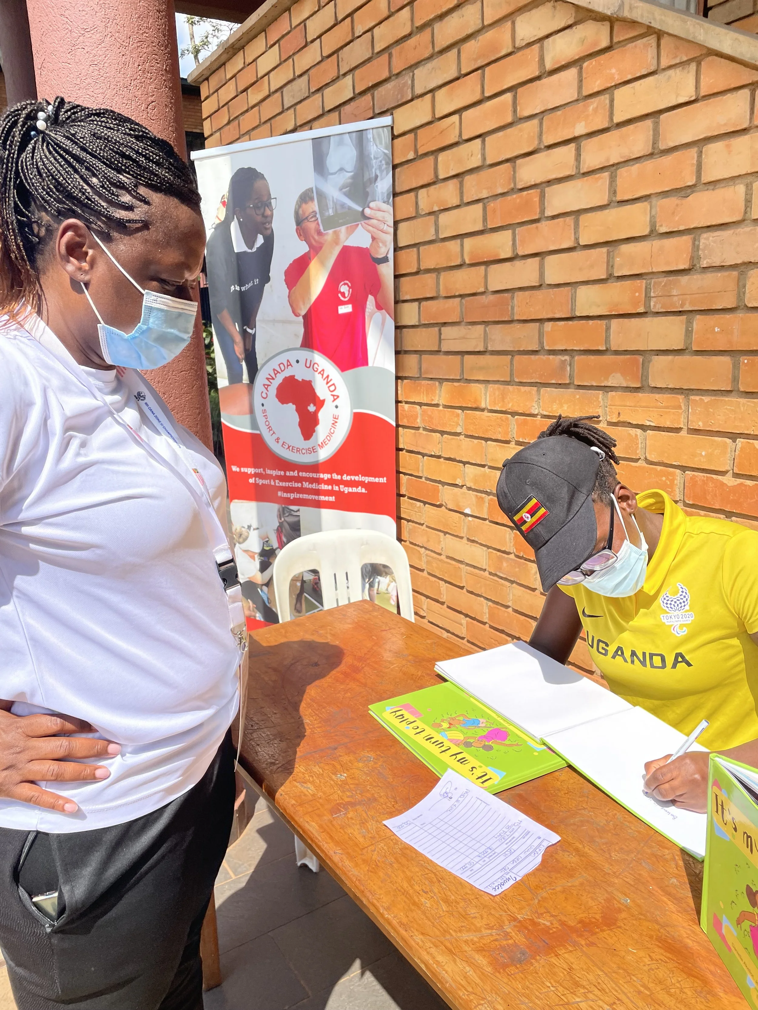 A woman in a white shirt and face mask is standing at a desk while a young man in a yellow Uganda Olympic team shirt, cap, and face mask is sitting and writing in a book. There is a banner in the background that supports sports and exercise medicine development in Uganda.