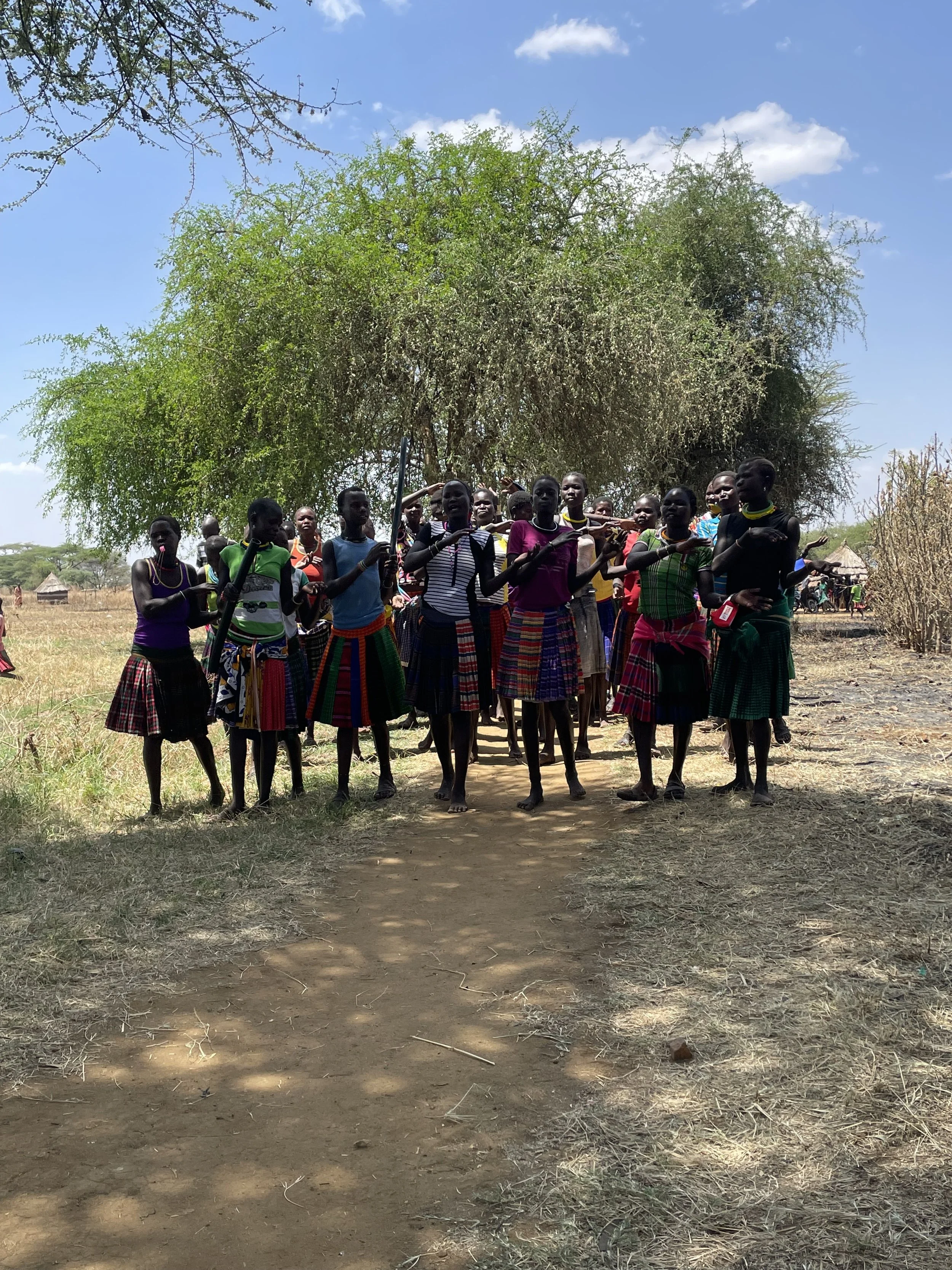 Group of people dressed in traditional clothing standing on a dirt path under a large tree in Karamoja Uganda.