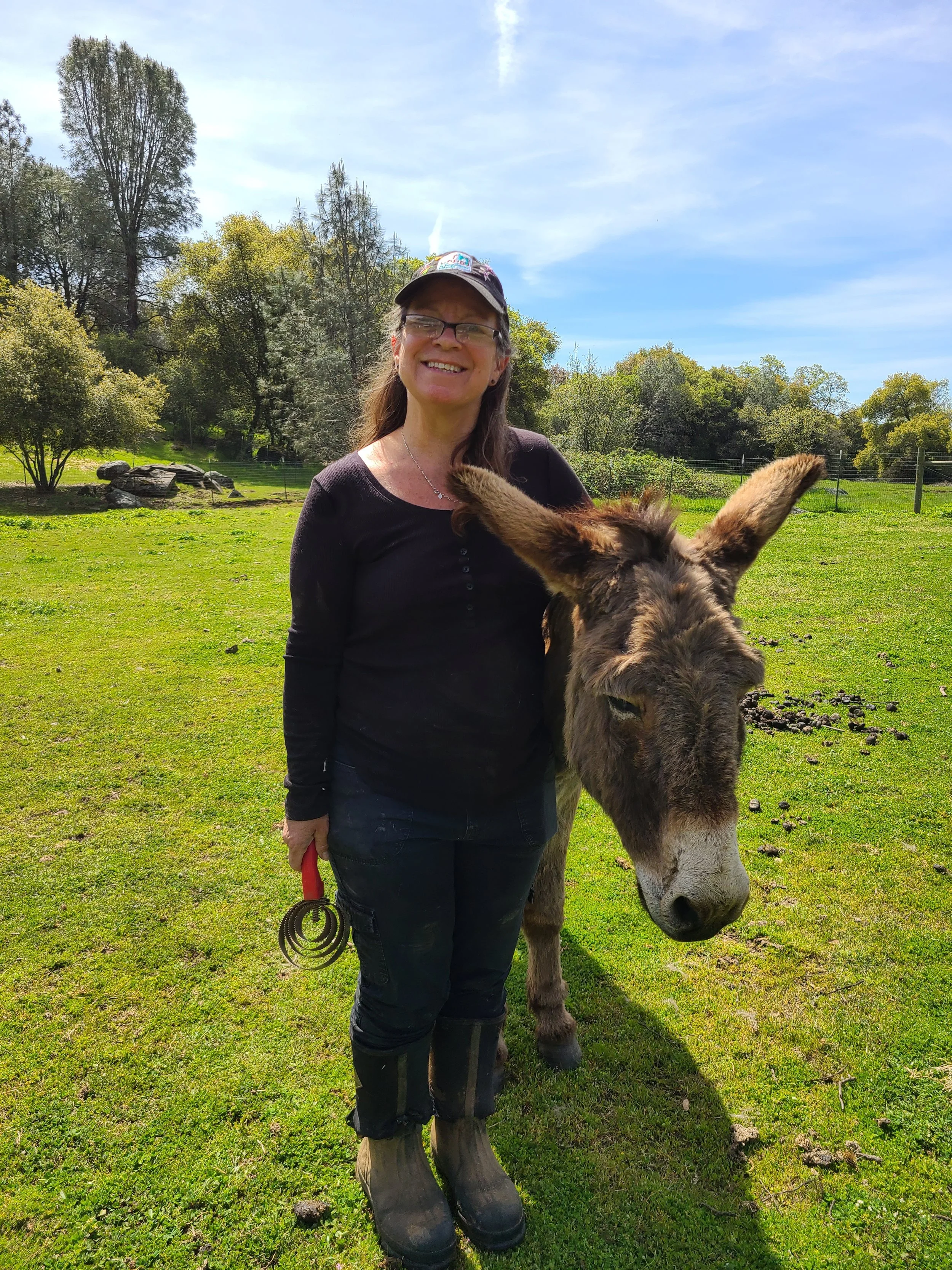 A woman standing on a lush green field, smiling, with a donkey beside her. The woman is dressed in black clothes, wearing boots, a cap, and glasses. The background includes trees and a partly cloudy blue sky.