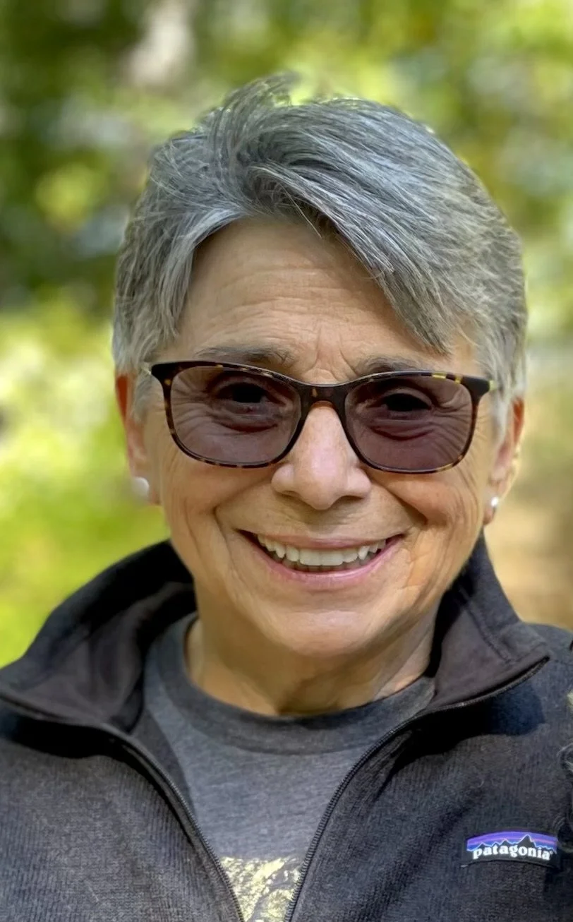 Smiling elderly woman with short gray hair wearing sunglasses and a black Patagonia jacket outdoors.