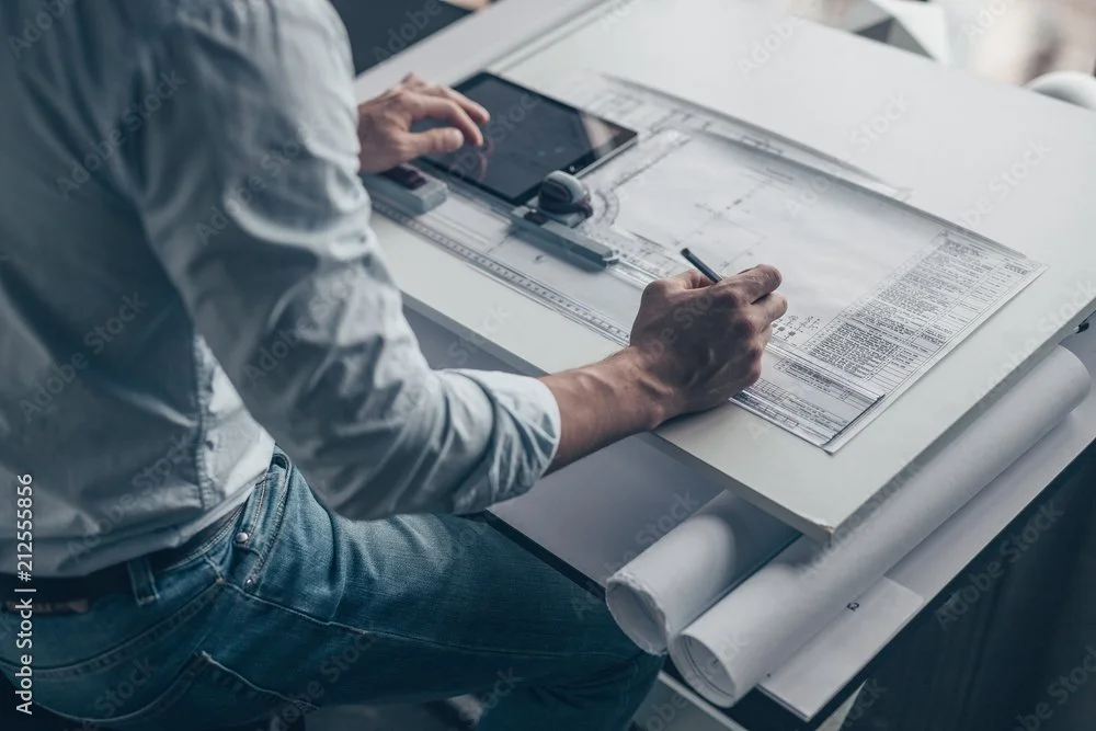 A person working on architectural or engineering plans at a drafting table with a tablet and drafting tools.