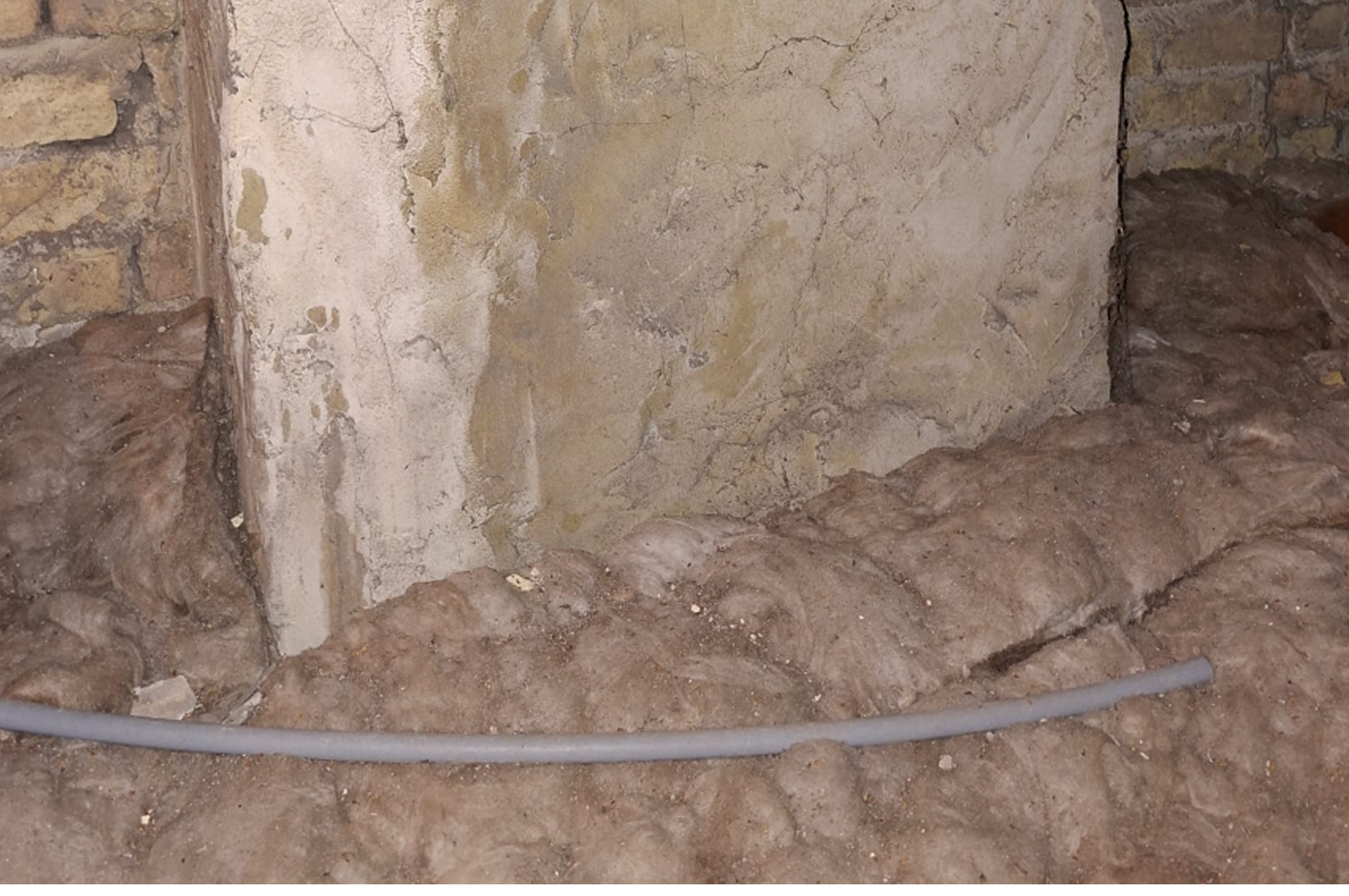 A construction site showing a dirt floor, a concrete wall with visible cracks, and a gray electrical conduit pipe on the ground.