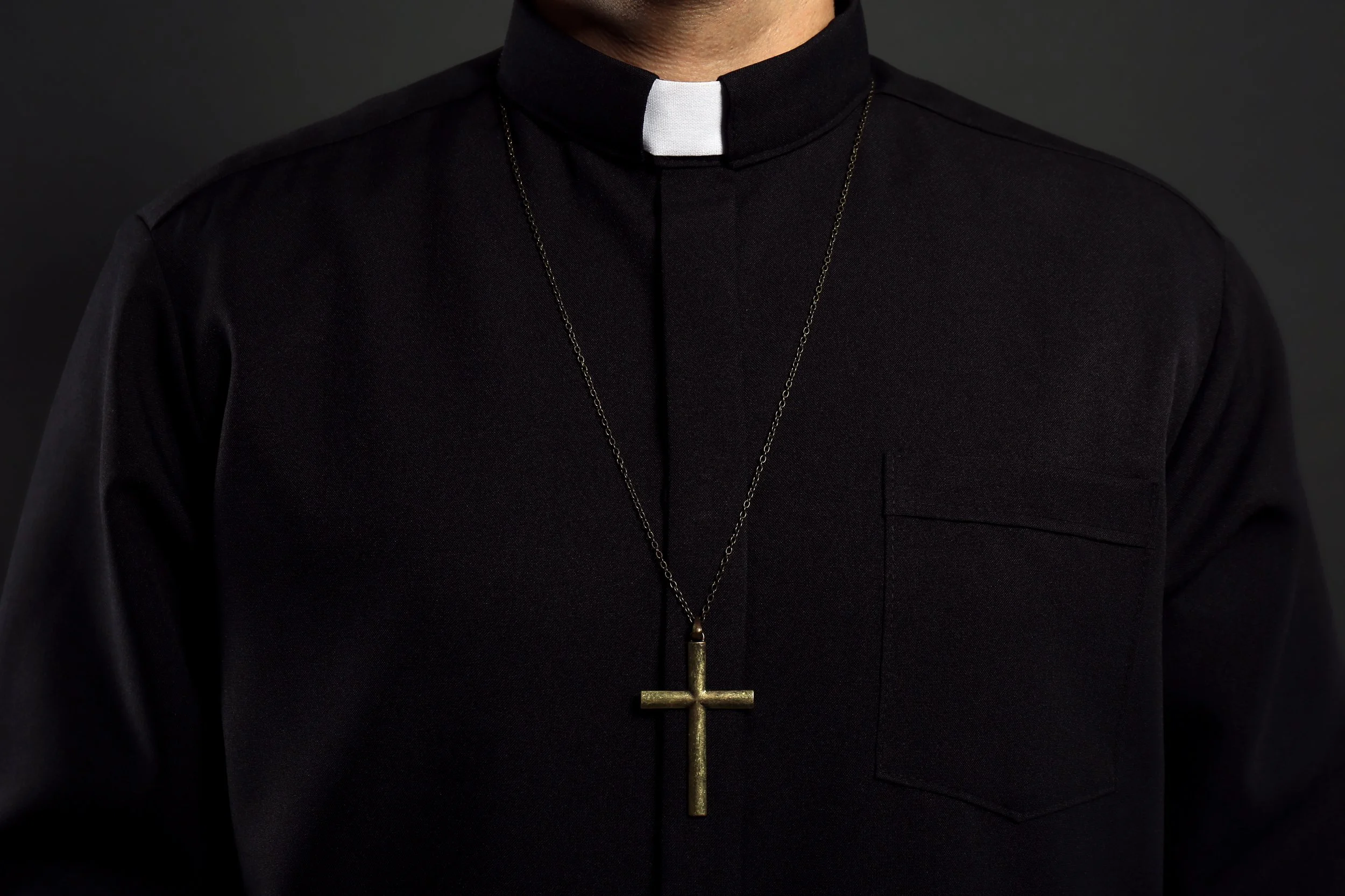 Close-up of a priest wearing a black clerical shirt, white collar, and a gold cross necklace.
